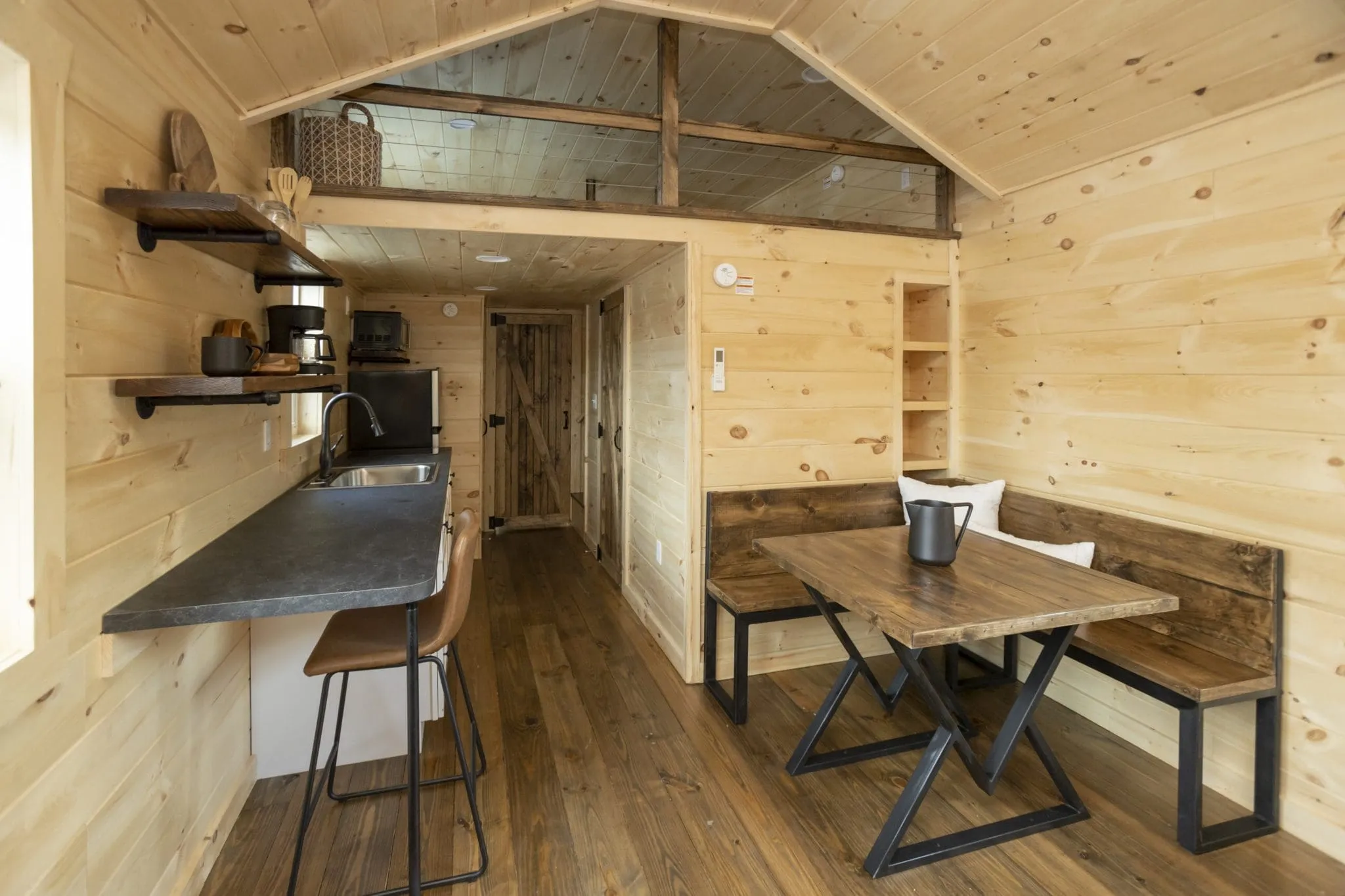 Compact wooden cabin interior with a small kitchenette, bar stool, corner bench with cushions, and a wooden table holding a black pitcher.