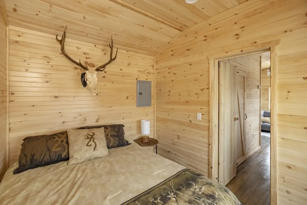 Cozy wooden bedroom with a bed featuring deer-themed pillows and blanket, a mounted antler skull on the wall, and a doorway leading to a hallway.