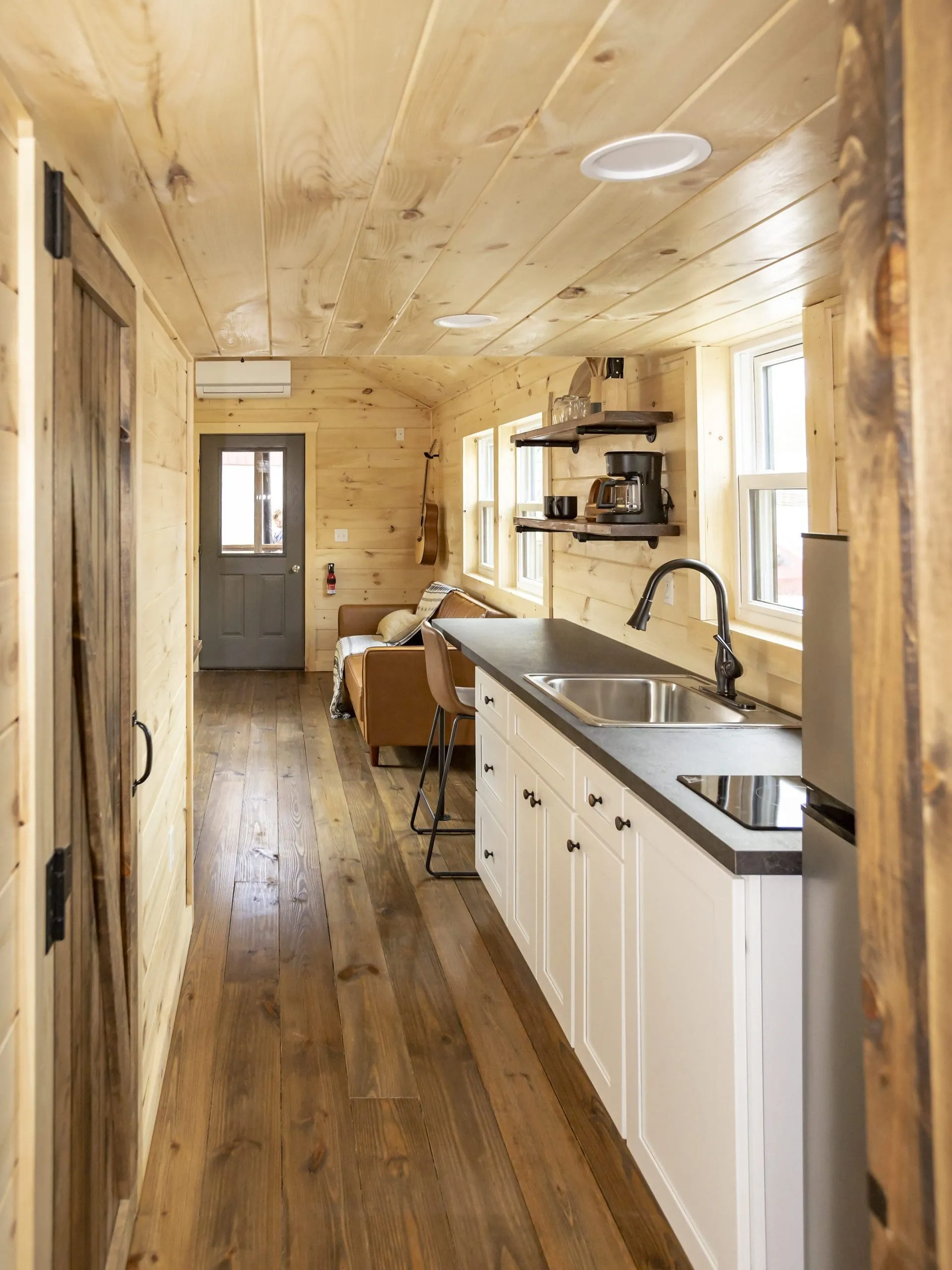 Interior of a narrow modern tiny cabin with wood-paneled walls and ceiling, white kitchen cabinets, black countertop, stainless steel sink and refrigerator, and a brown leather couch.