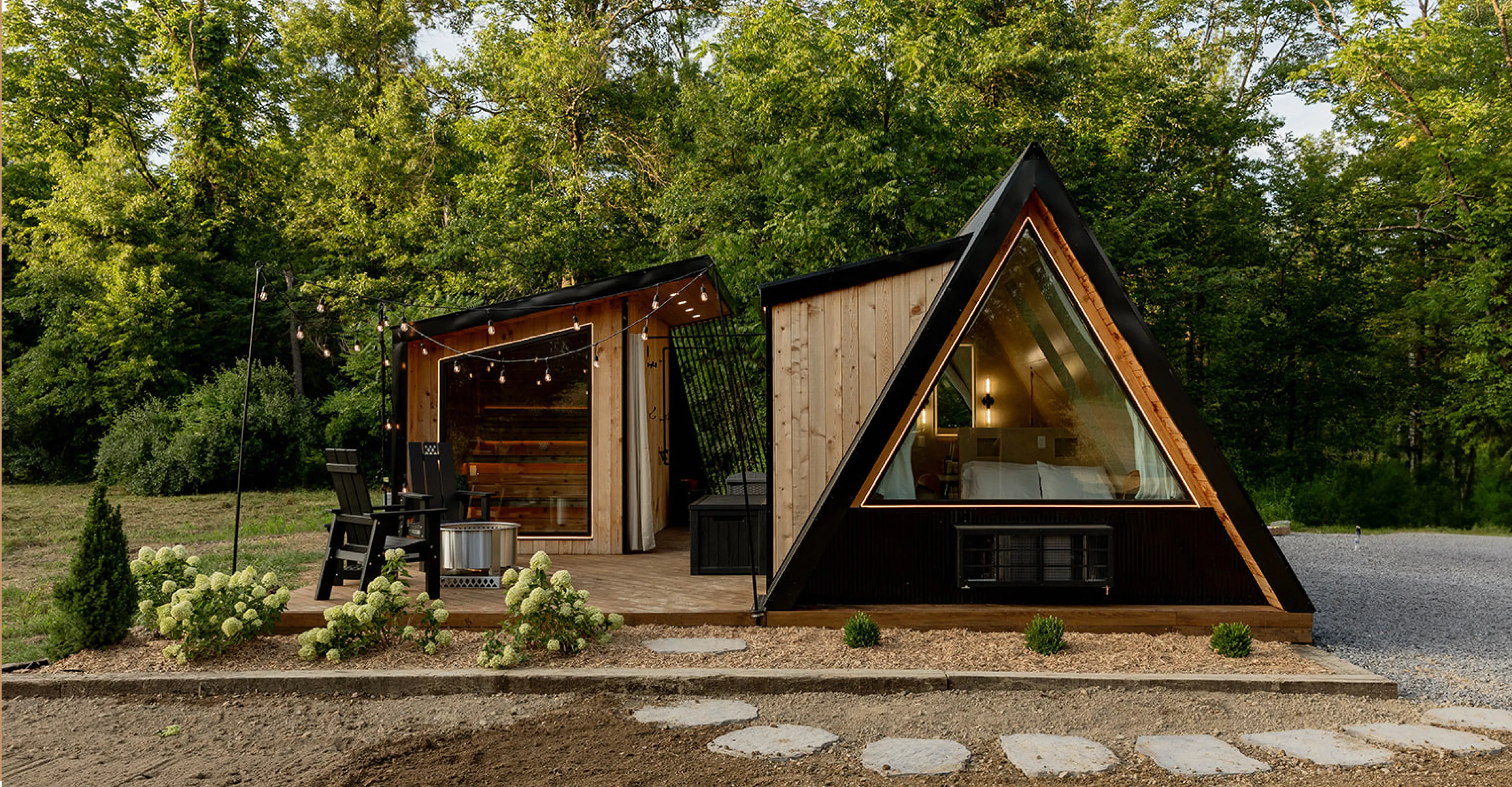 Modern wooden A-frame cabin with a triangular window, patio seating, string lights, and surrounded by greenery.