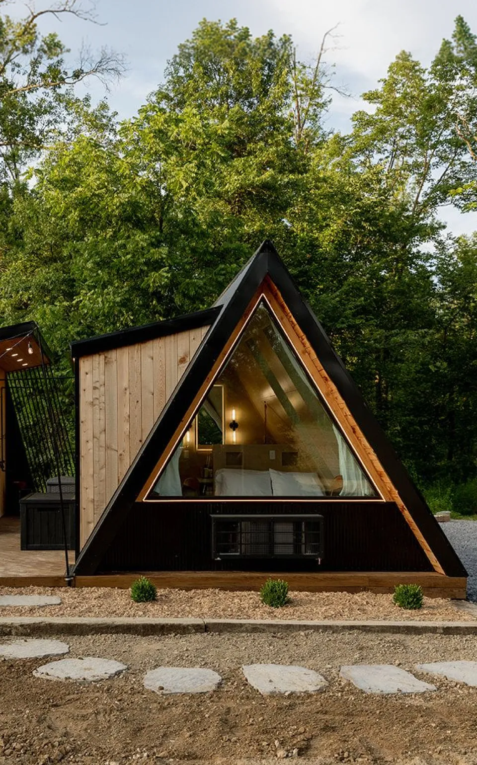 Modern wooden A-frame cabin with a triangular window, patio seating, string lights, and surrounded by greenery.