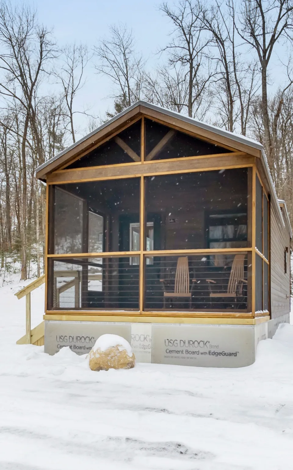 Small wooden cabin with screened porch surrounded by snow and leafless trees in winter.
