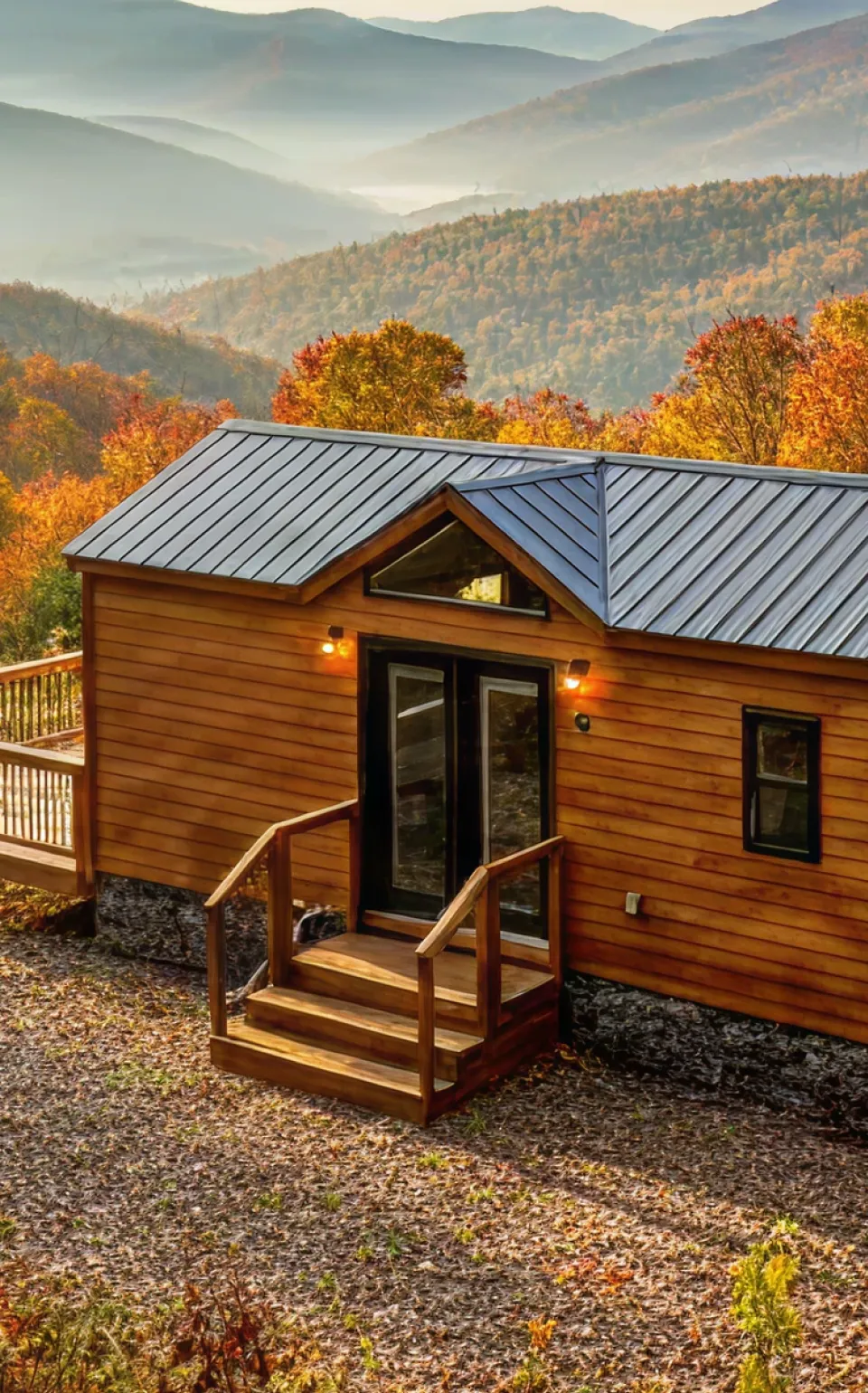 Small wooden cabin with a metal roof and a deck surrounded by autumn trees in a mountainous forest.