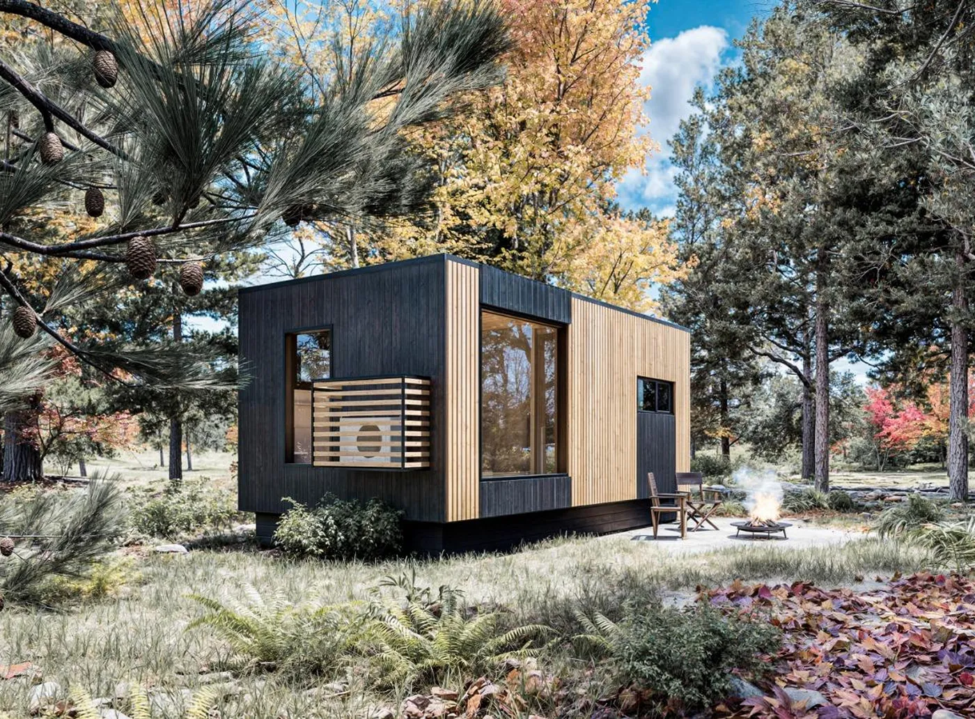A modern Vista model cabin with wood and dark paneling sits in a forest clearing surrounded by trees, with two chairs by a small fire pit in the foreground.