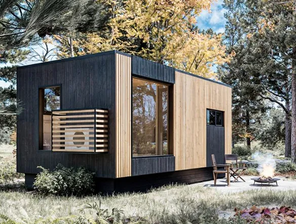 A modern tiny house with wood and dark paneling sits in a forest clearing surrounded by trees, with two chairs by a small fire pit in the foreground.