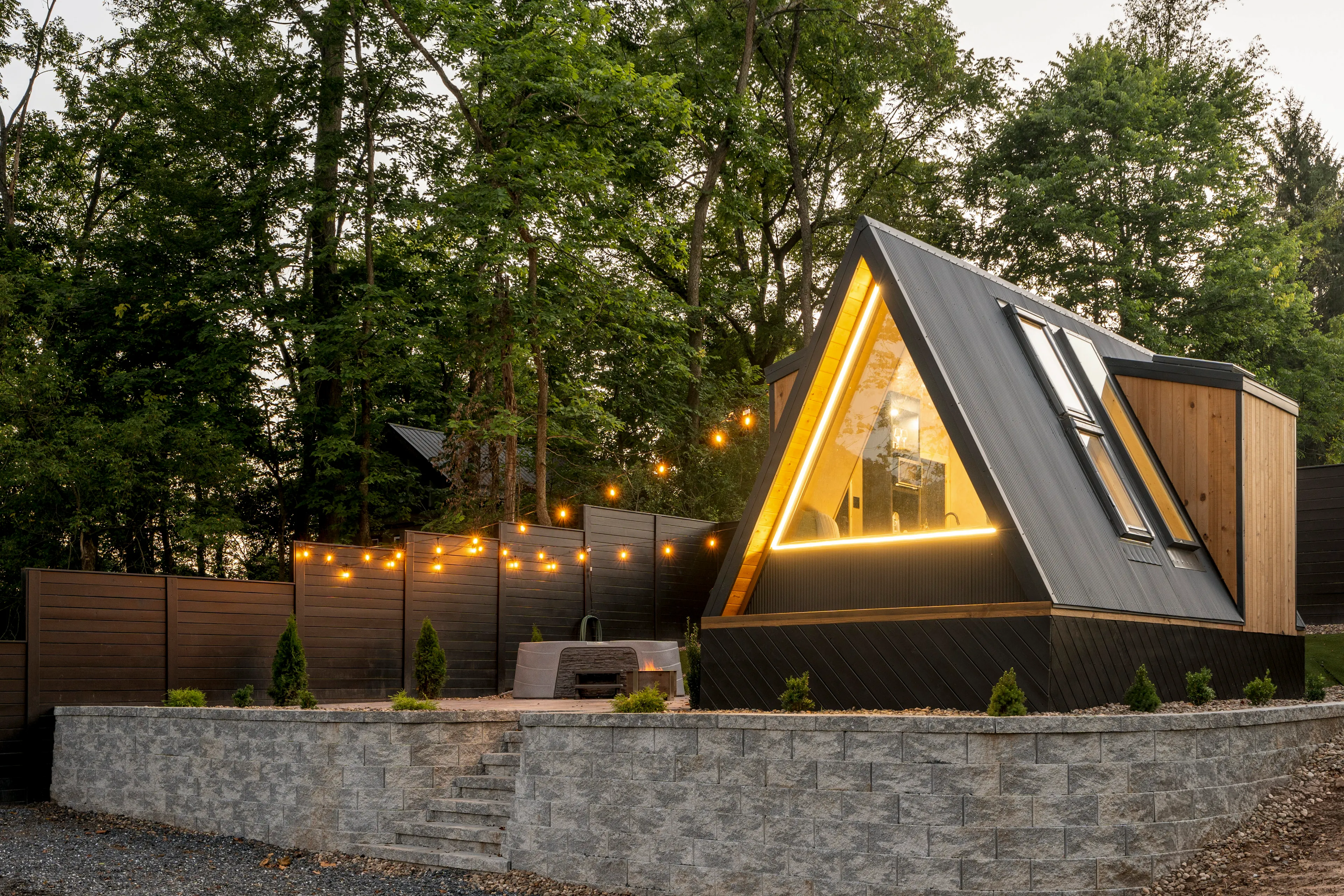 A modern A-frame cabin with large windows and string lights is set on a raised stone platform, surrounded by trees and wooden fencing.