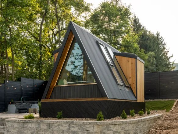 A modern A-frame cabin with large windows and string lights is set on a raised stone platform, surrounded by trees and wooden fencing.