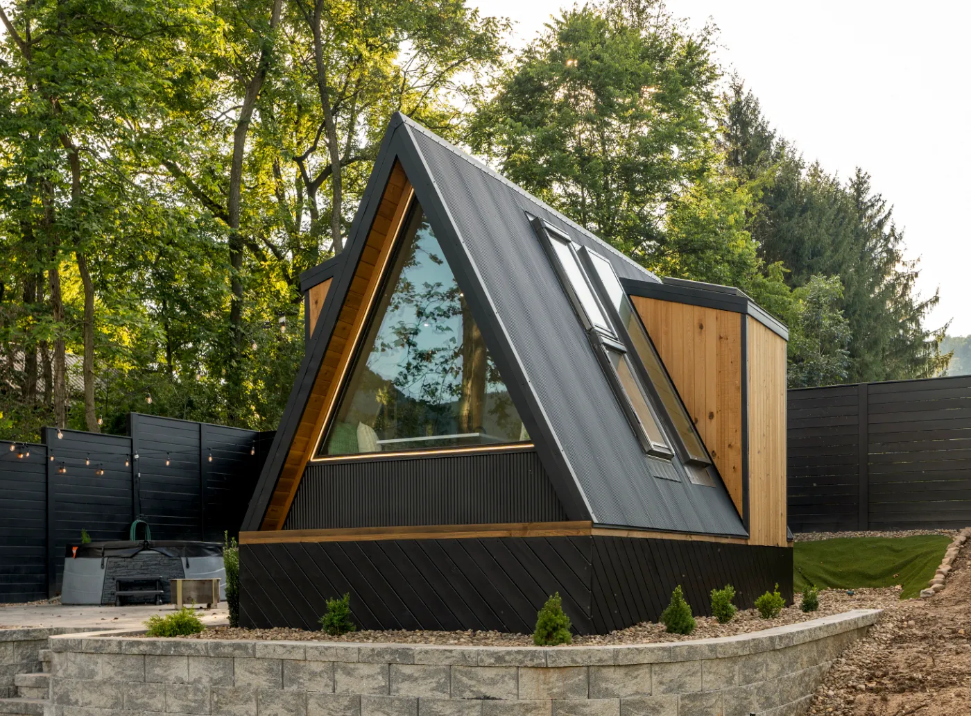 A modern A-frame cabin with large windows and string lights is set on a raised stone platform, surrounded by trees and wooden fencing.