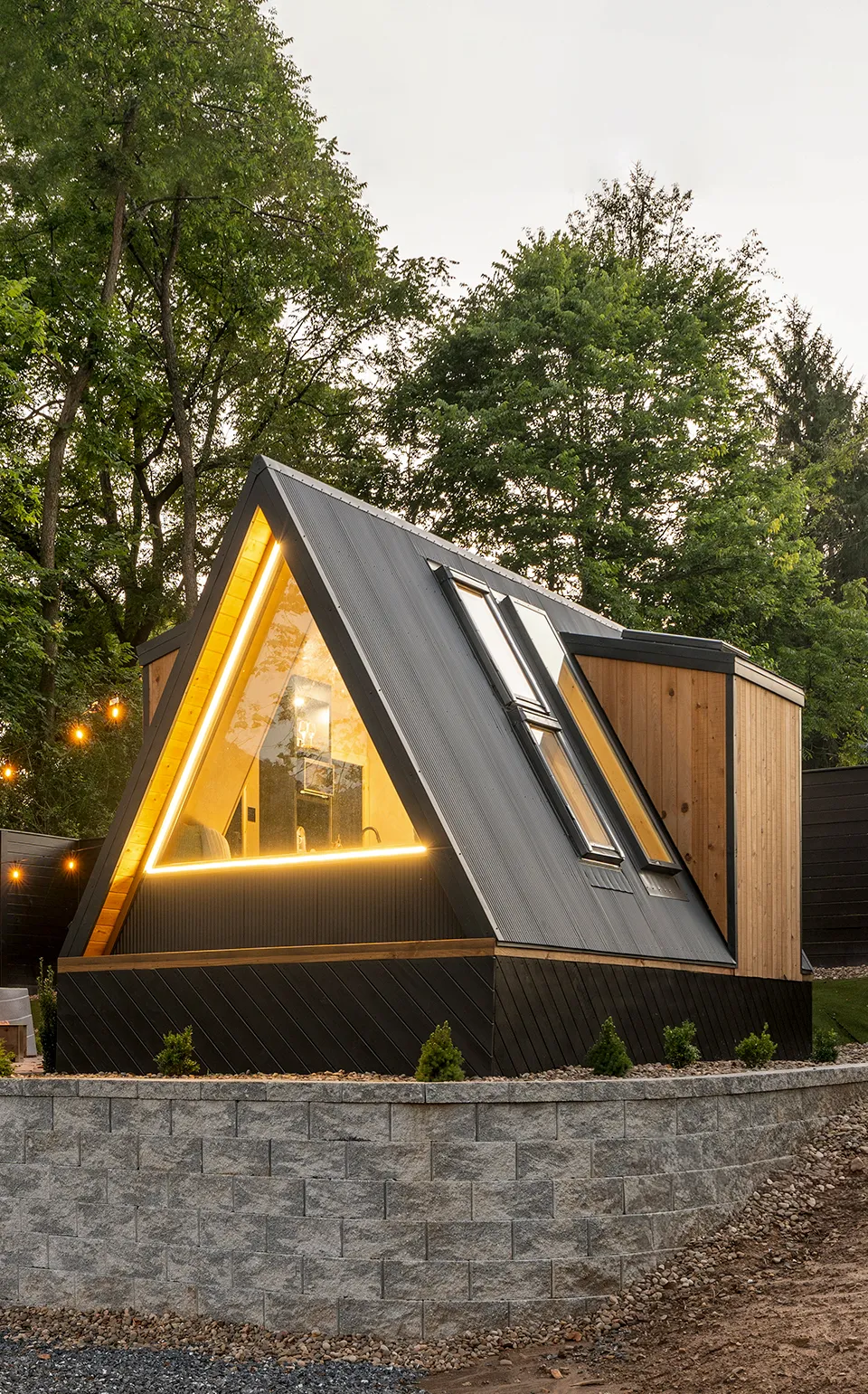 A modern A-frame cabin with large windows and string lights is set on a raised stone platform, surrounded by trees and wooden fencing.