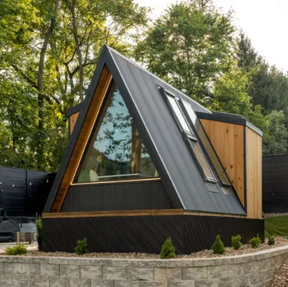 The Wanderhut Deluxe A-frame cabin with a black metal roof, large triangular window, and wood-paneled side, set behind a stone retaining wall.