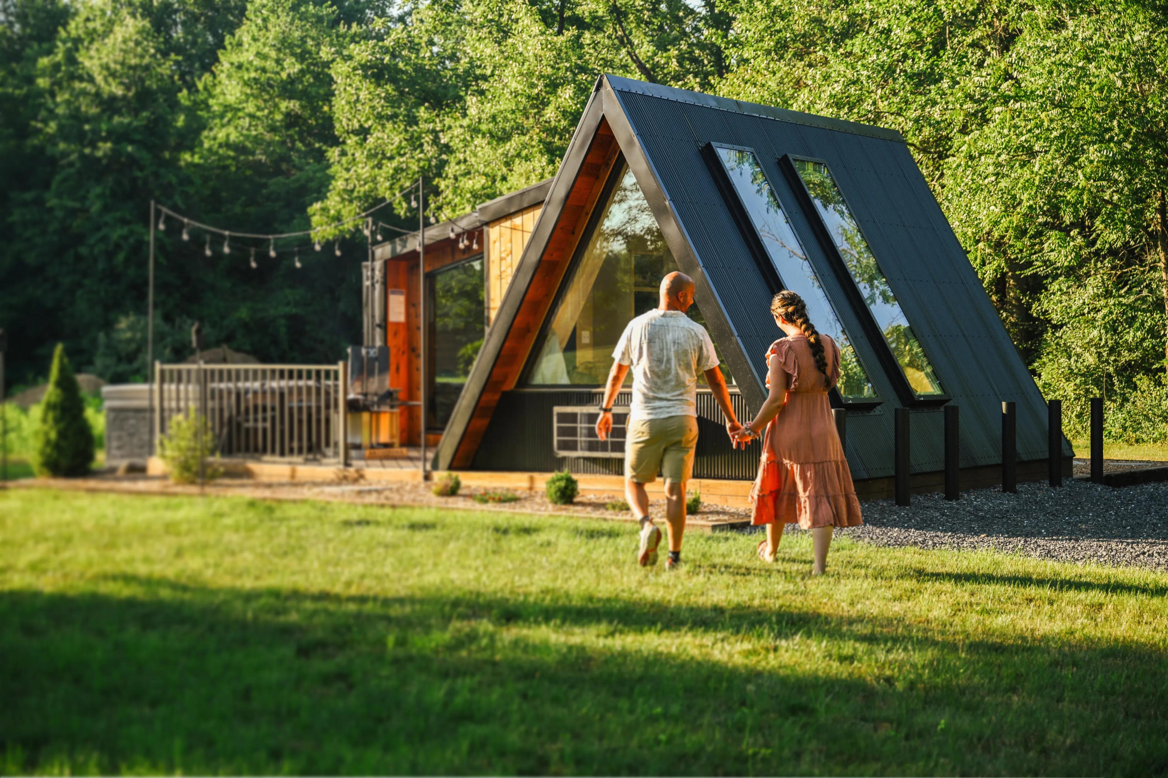 A couple walks hand-in-hand toward an A-frame cabin in a wooded area.