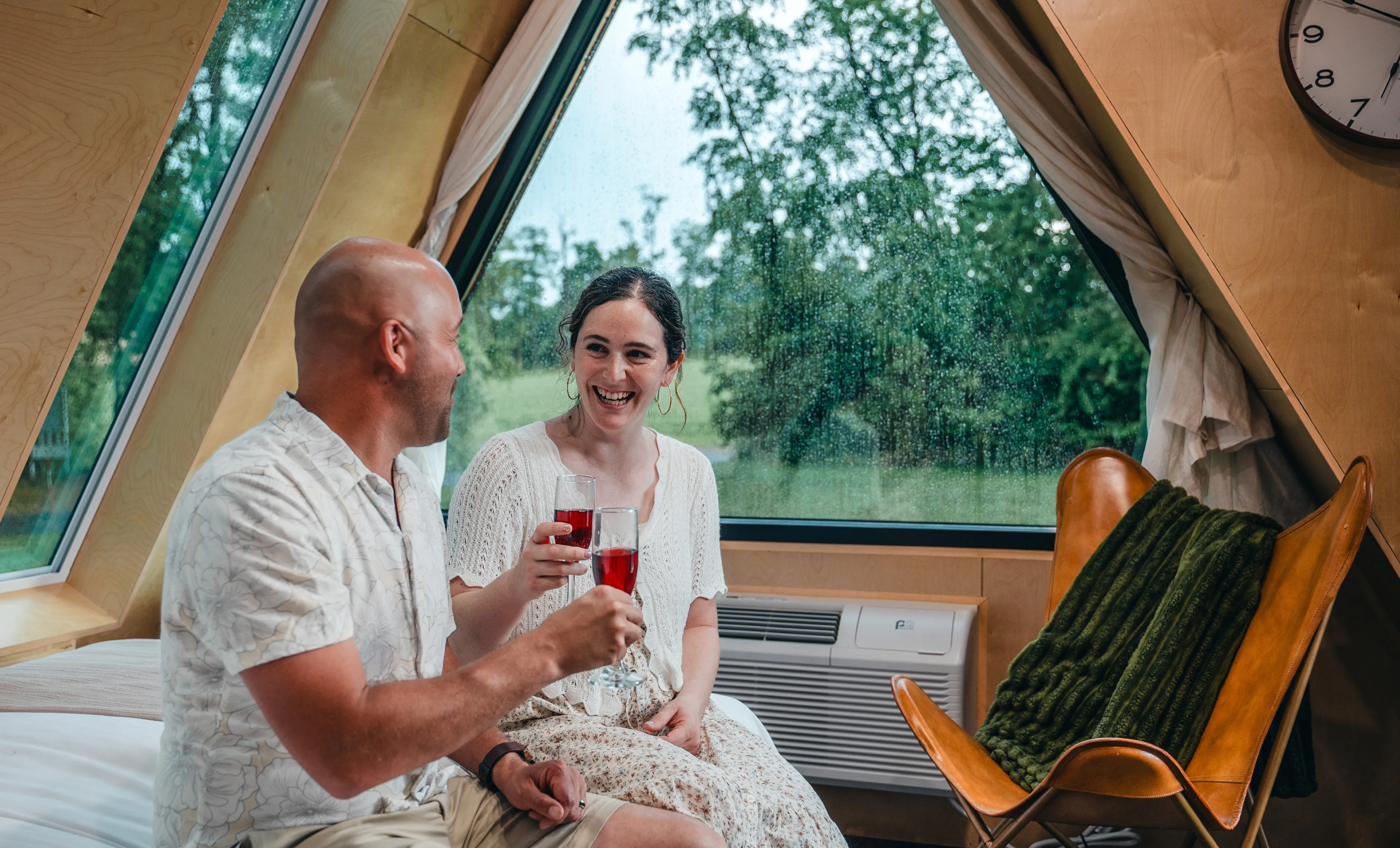 A couple sitting on a bed in an A-frame cabin cheers with glasses of red wine in front of a window.