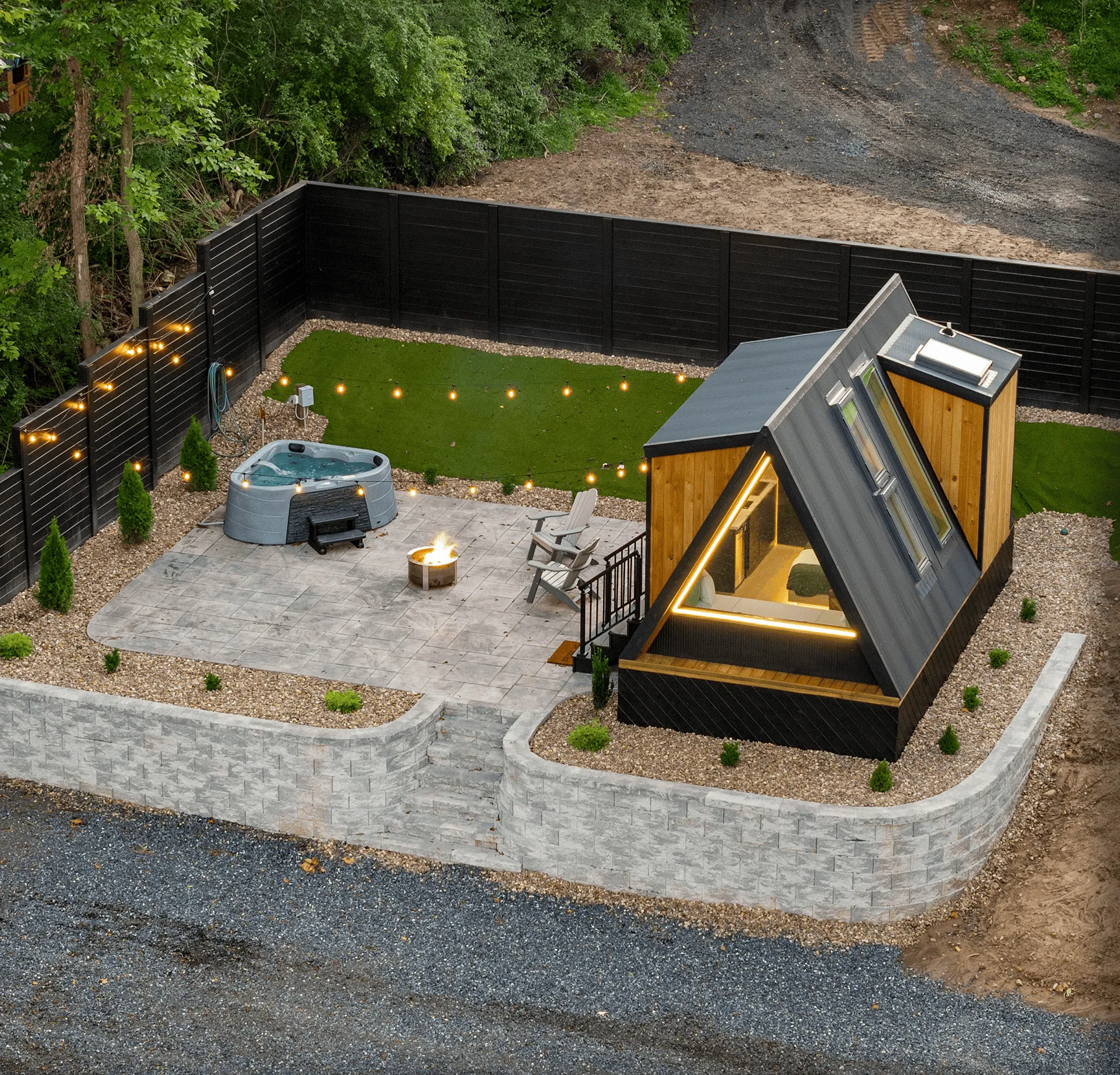 A small A-frame cabin with a patio featuring a hot tub, fire pit, and two chairs, enclosed by a black fence.