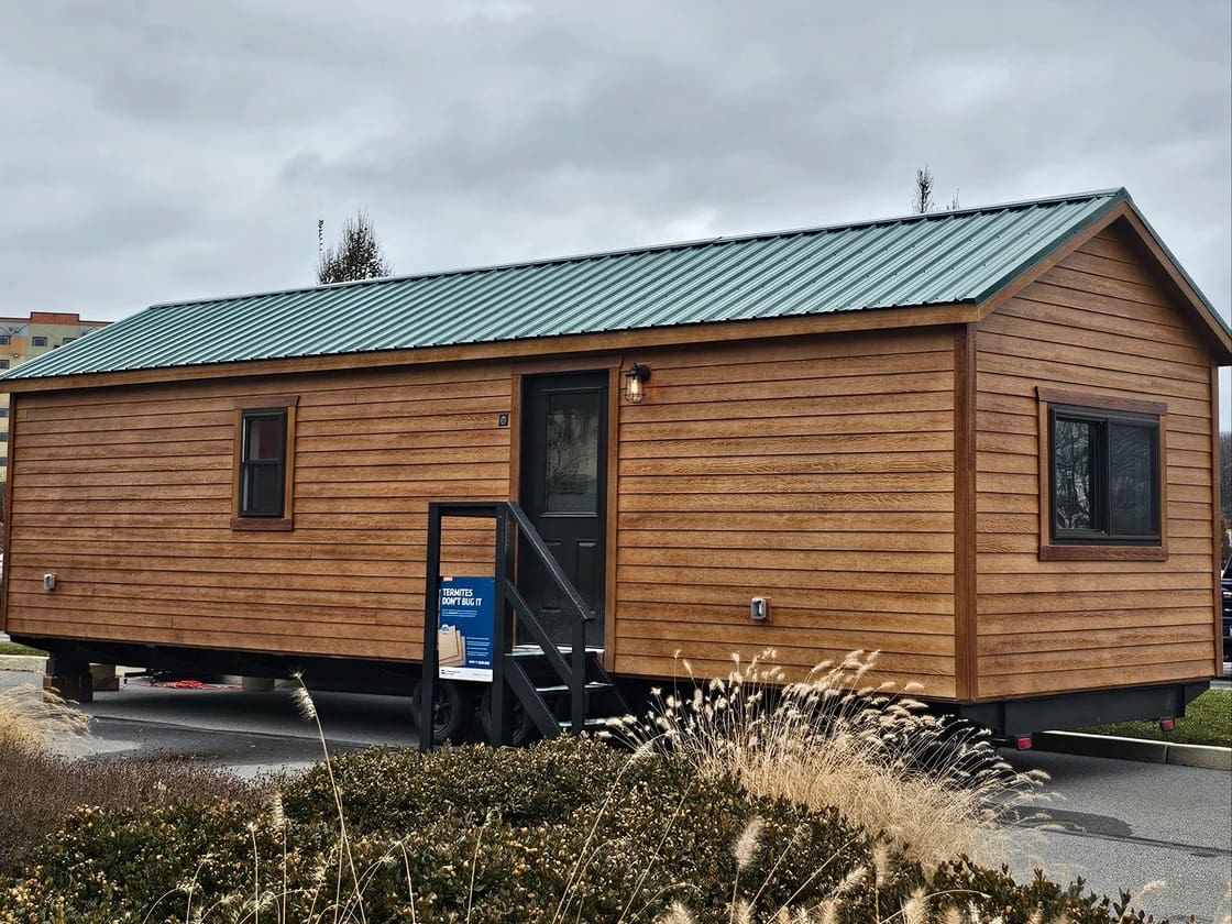 A small wooden house with a green metal roof sits on a trailer in an outdoor setting.