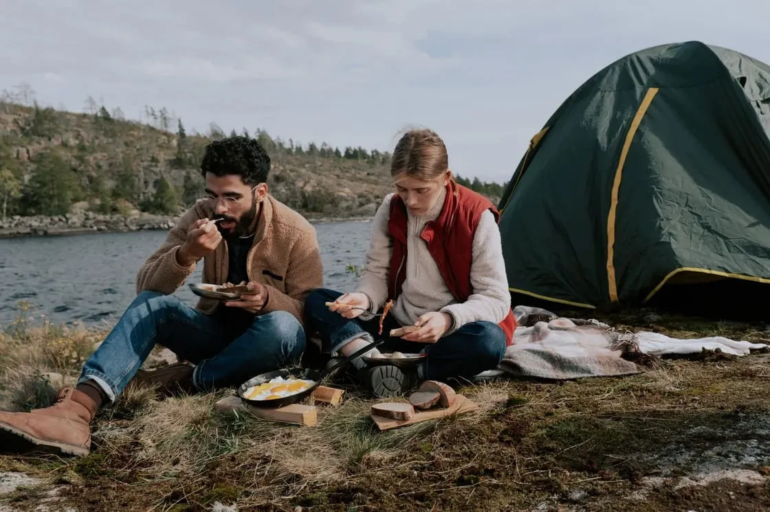 Two people sit on the grass by a lake, eating food beside a tent and camping gear, with trees and hills in the background.