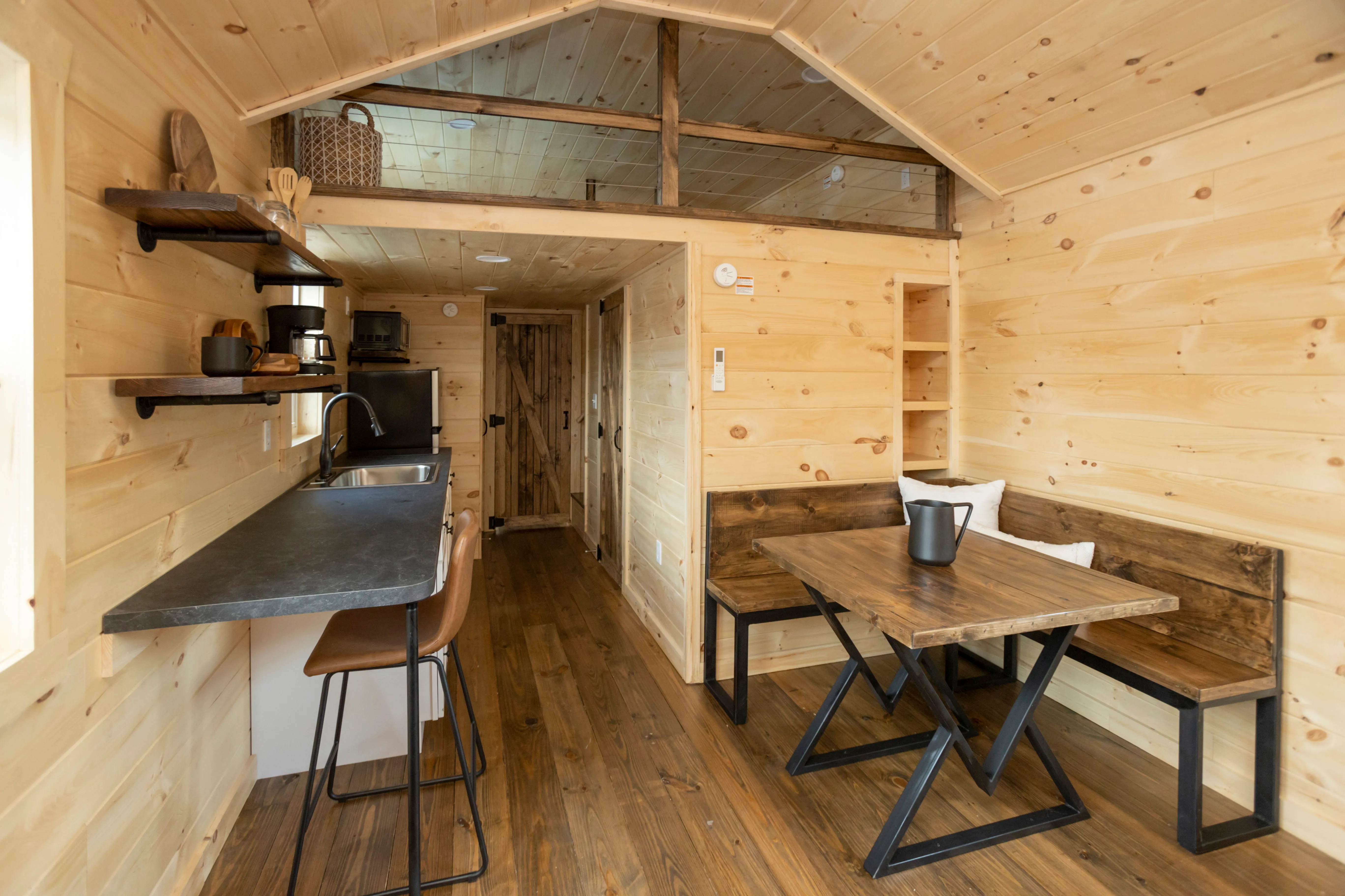 Interior of a small wooden cabin with a kitchenette, bar stools, a table with benches, open shelving, and light wood paneling throughout.
