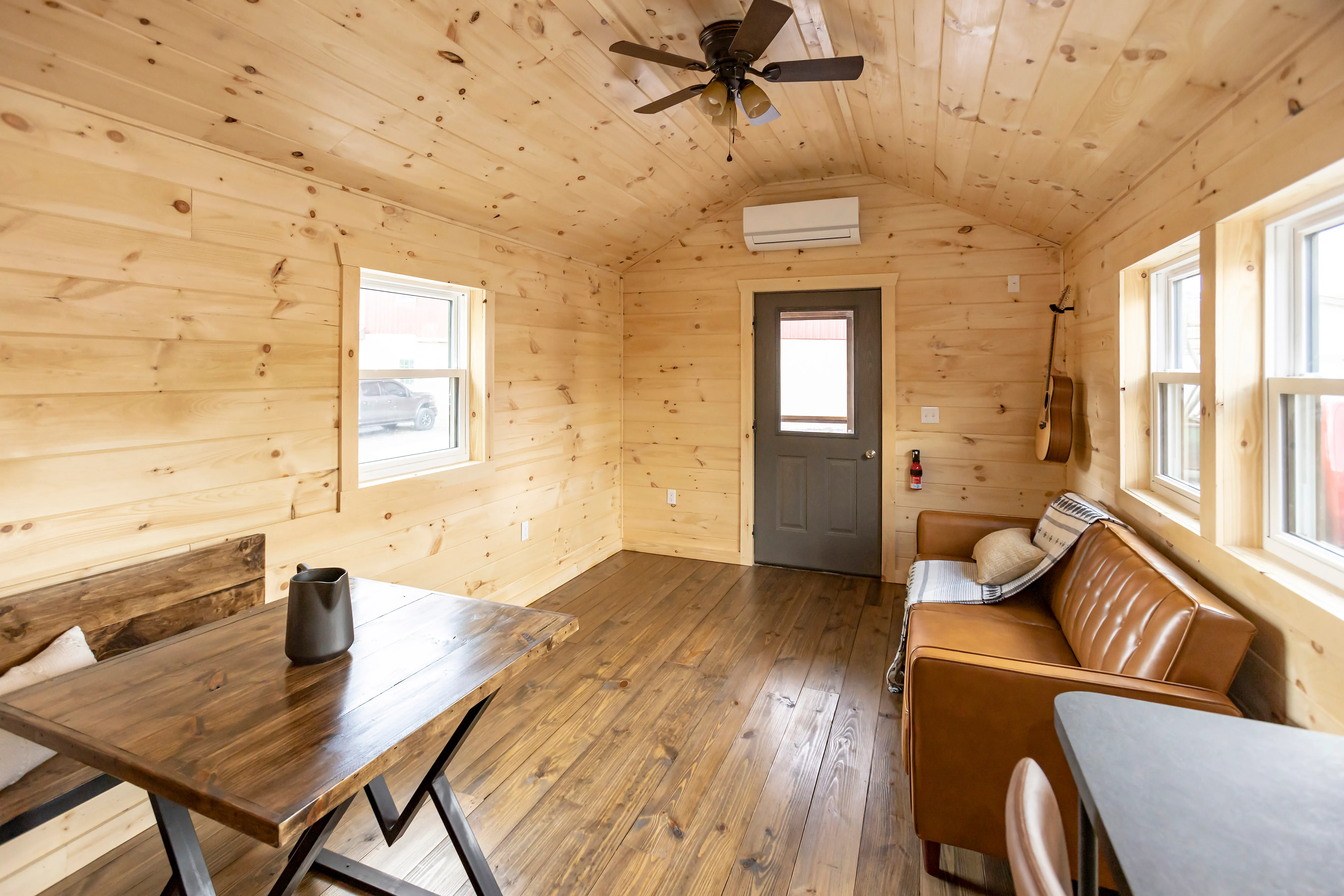 Small wooden cabin interior with a brown leather sofa, dining table, wood-paneled walls and ceiling, multiple windows, and a door.