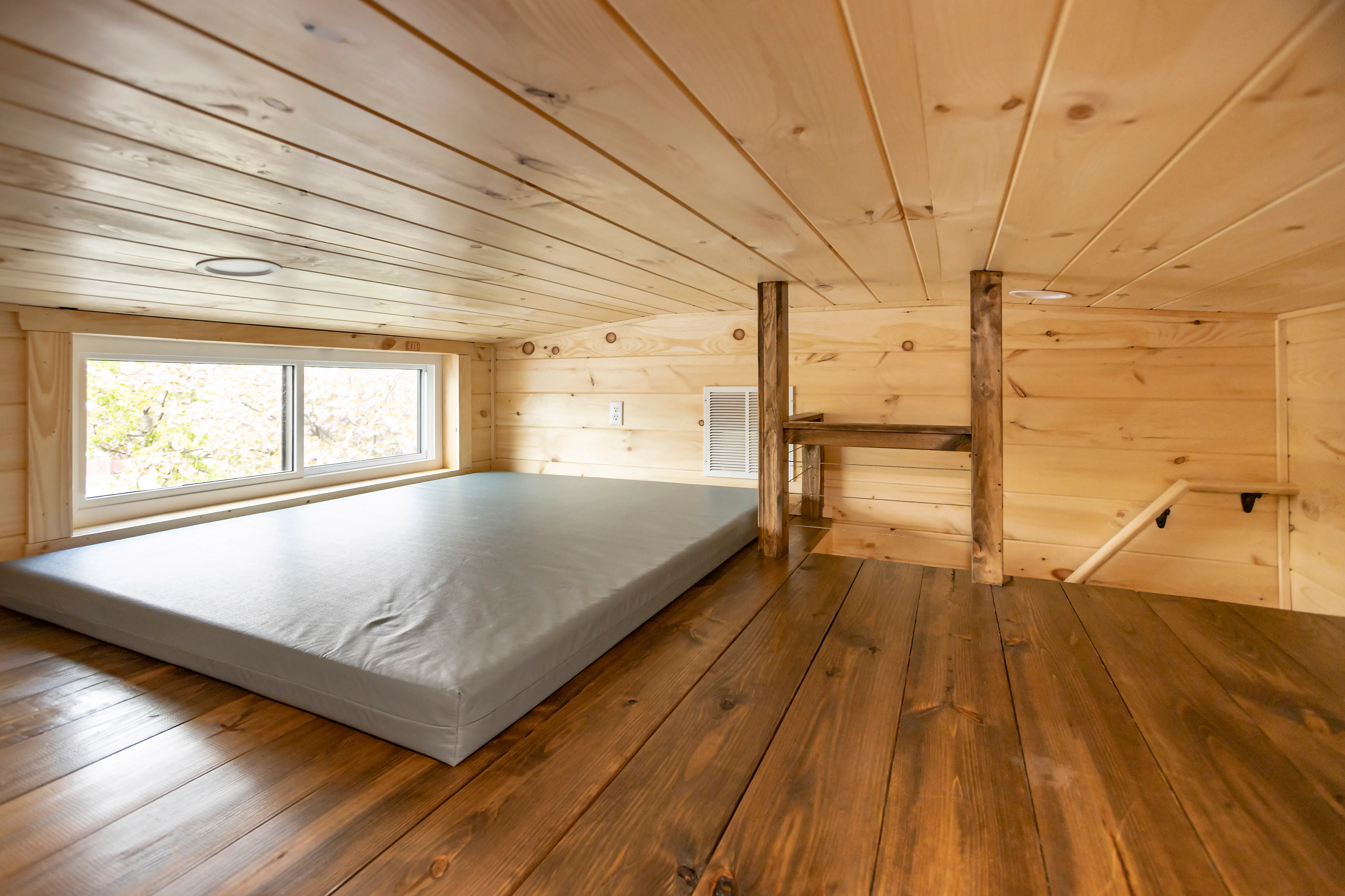 A small loft with wood-paneled walls and ceiling, a low mattress by a window, and wooden floors and posts.