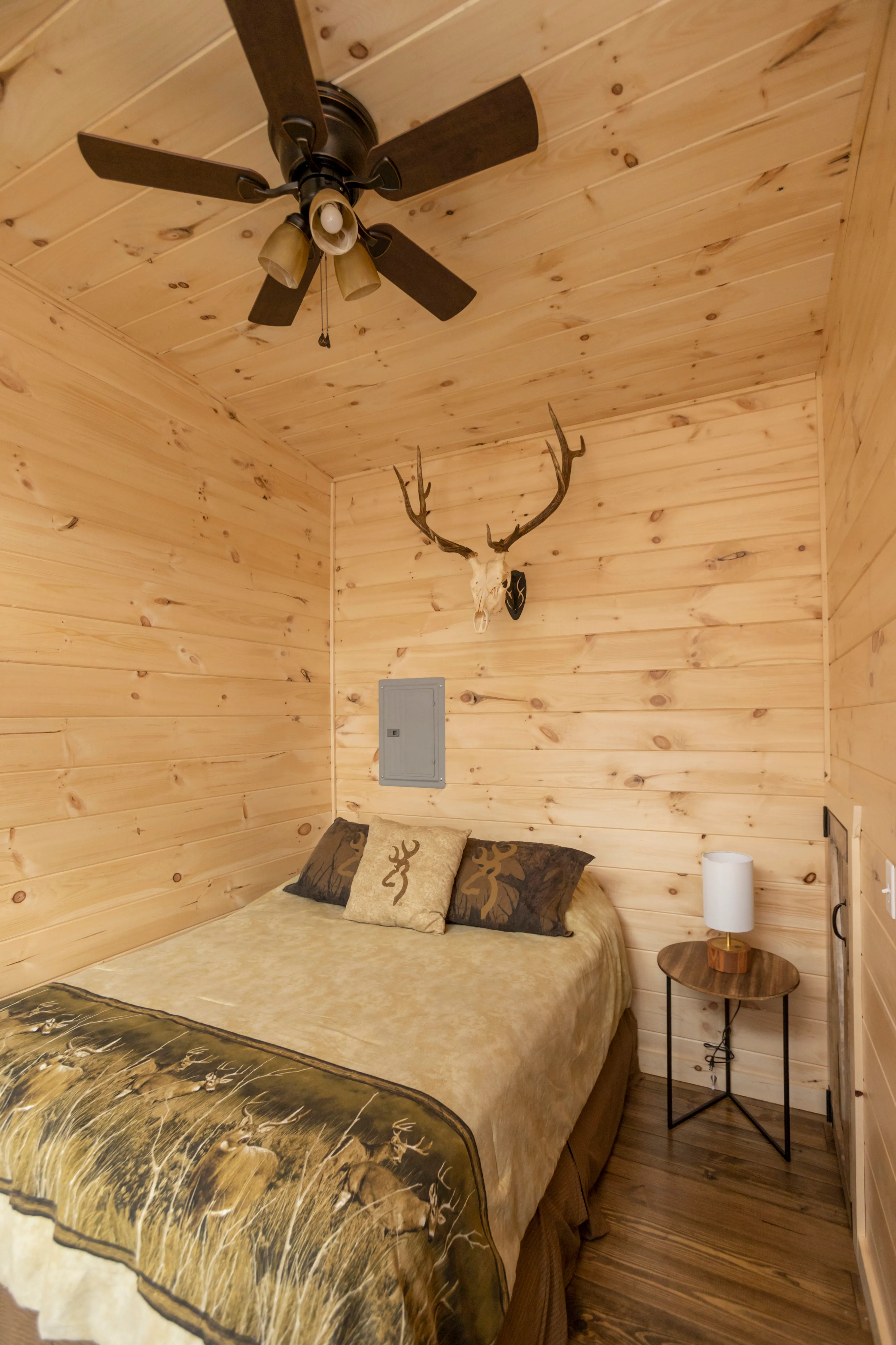 A small wooden room with a bed, a round side table with a lamp, ceiling fan, and mounted antlers on the wall above the bed.