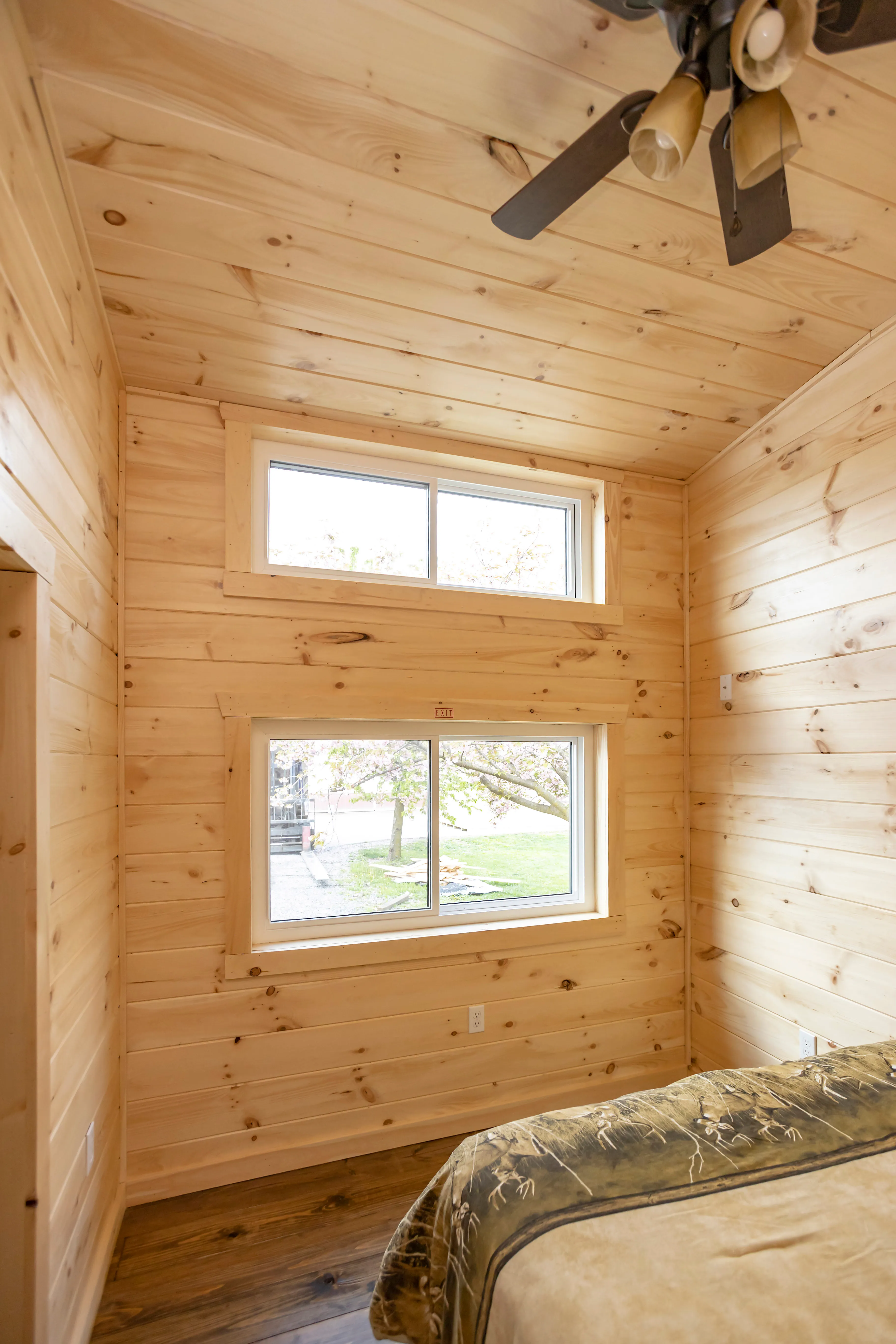 A small bedroom with light wood-paneled walls and ceiling, two rectangular windows, a ceiling fan, and a corner of a bed visible in the foreground.