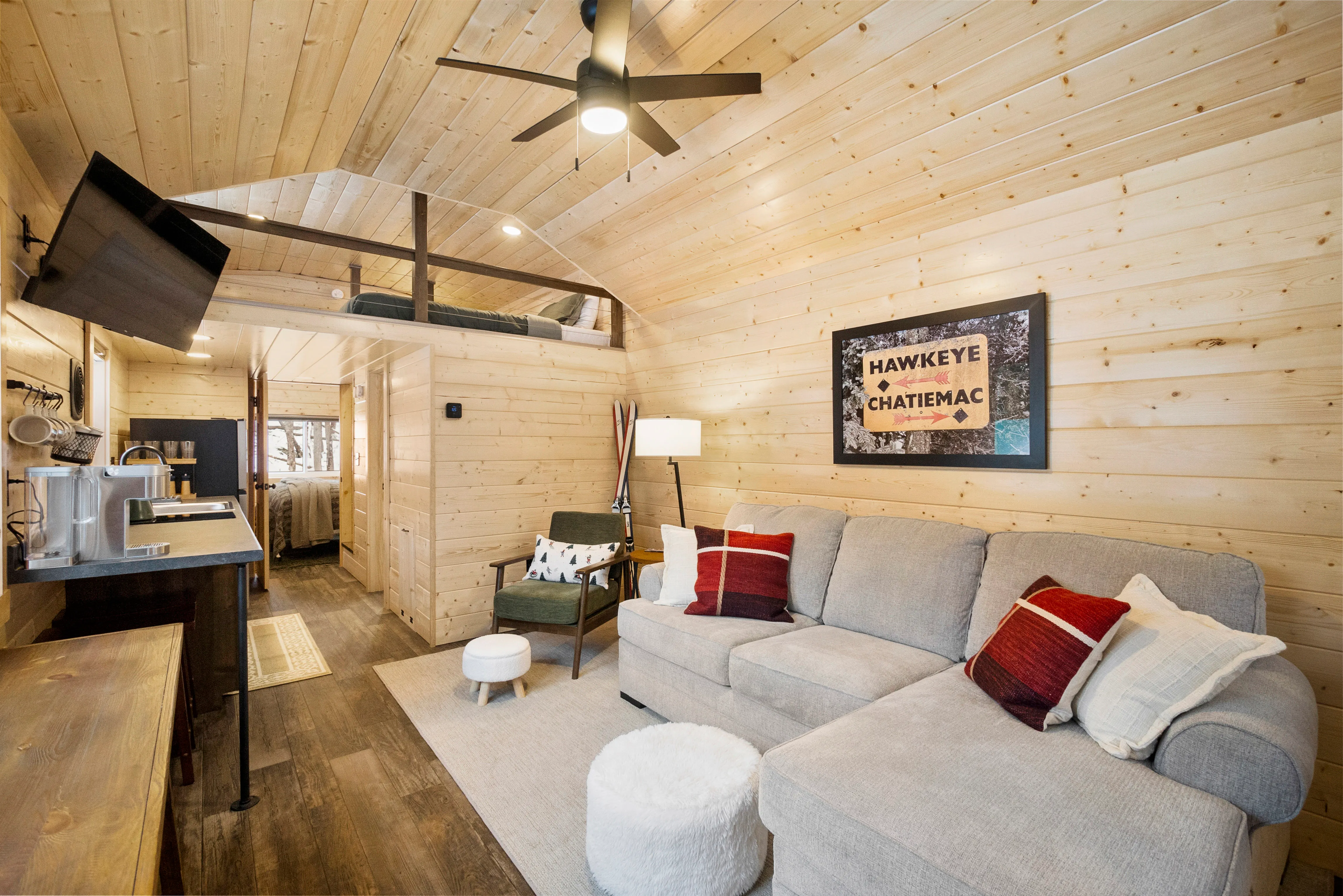A cozy living room in a wooden cabin features a gray sectional sofa, armchair, ottoman, wall art, TV, and ceiling fan, with a kitchen and loft area visible in the background.