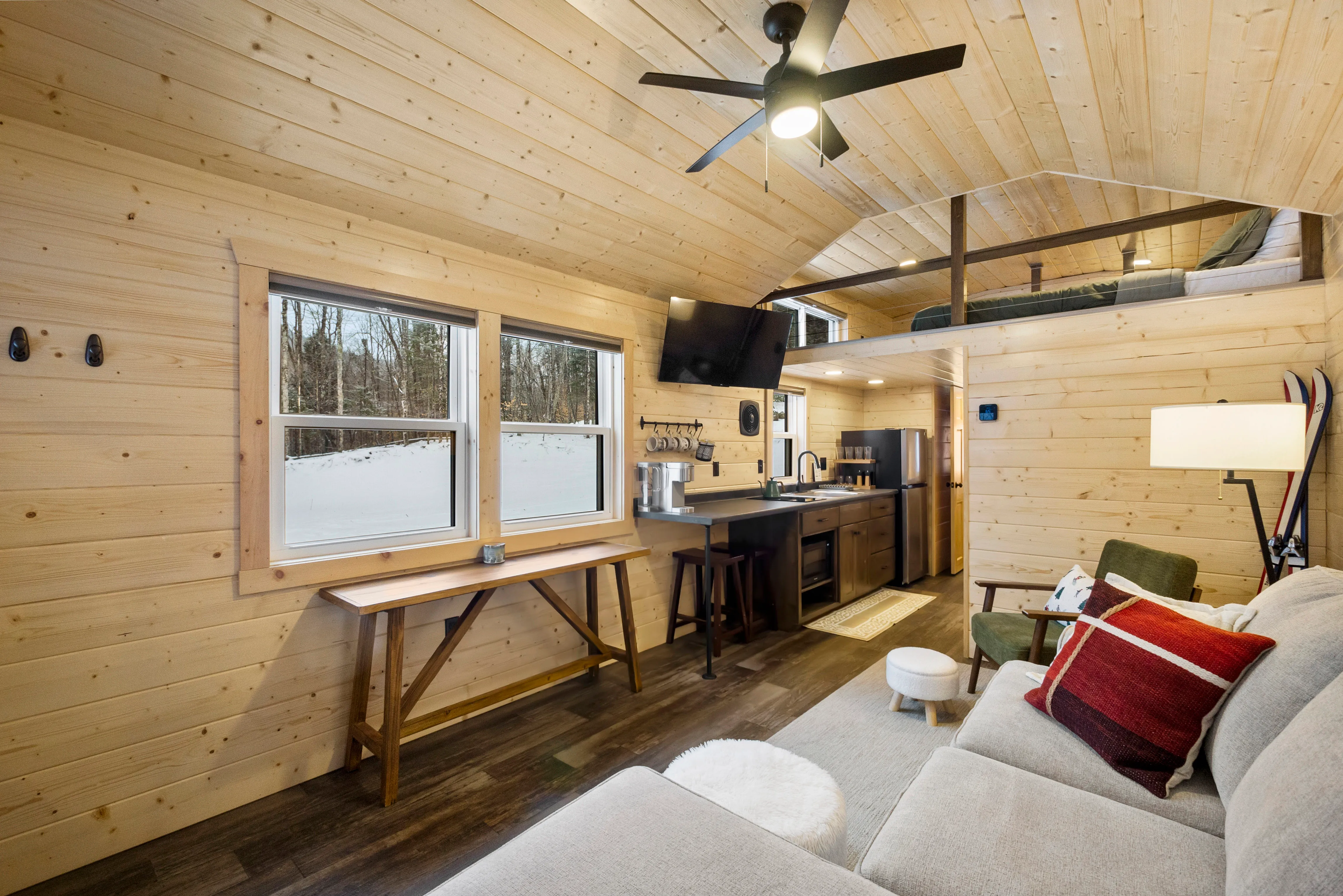 Modern tiny cabin interior with wood paneling, a loft bed, sofa, kitchenette, wall-mounted TV, dining table, and windows.
