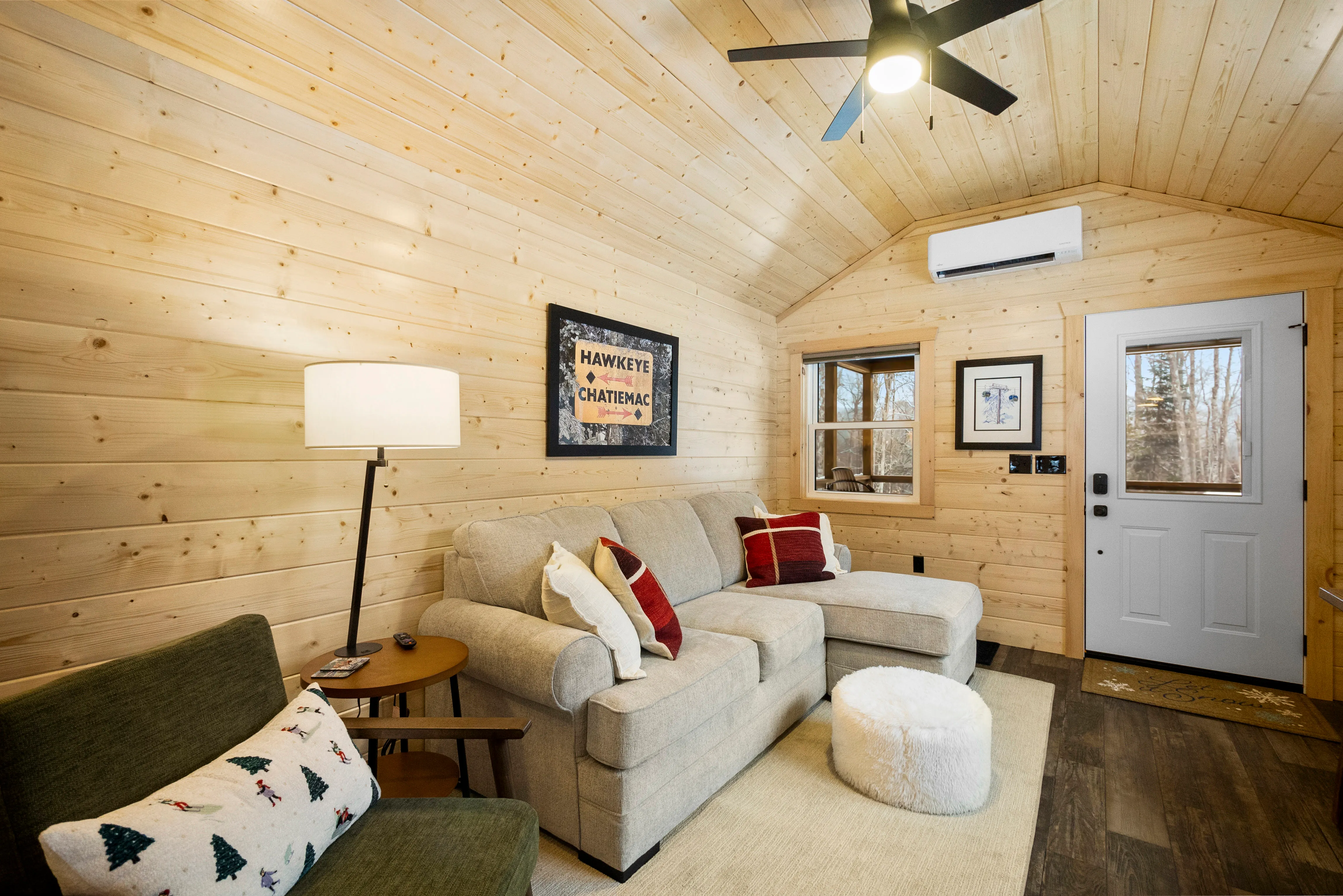 Cozy living room with wood-paneled walls, gray sectional sofa, green armchair, table lamp, framed art, ottoman, window, and a white door with a window.