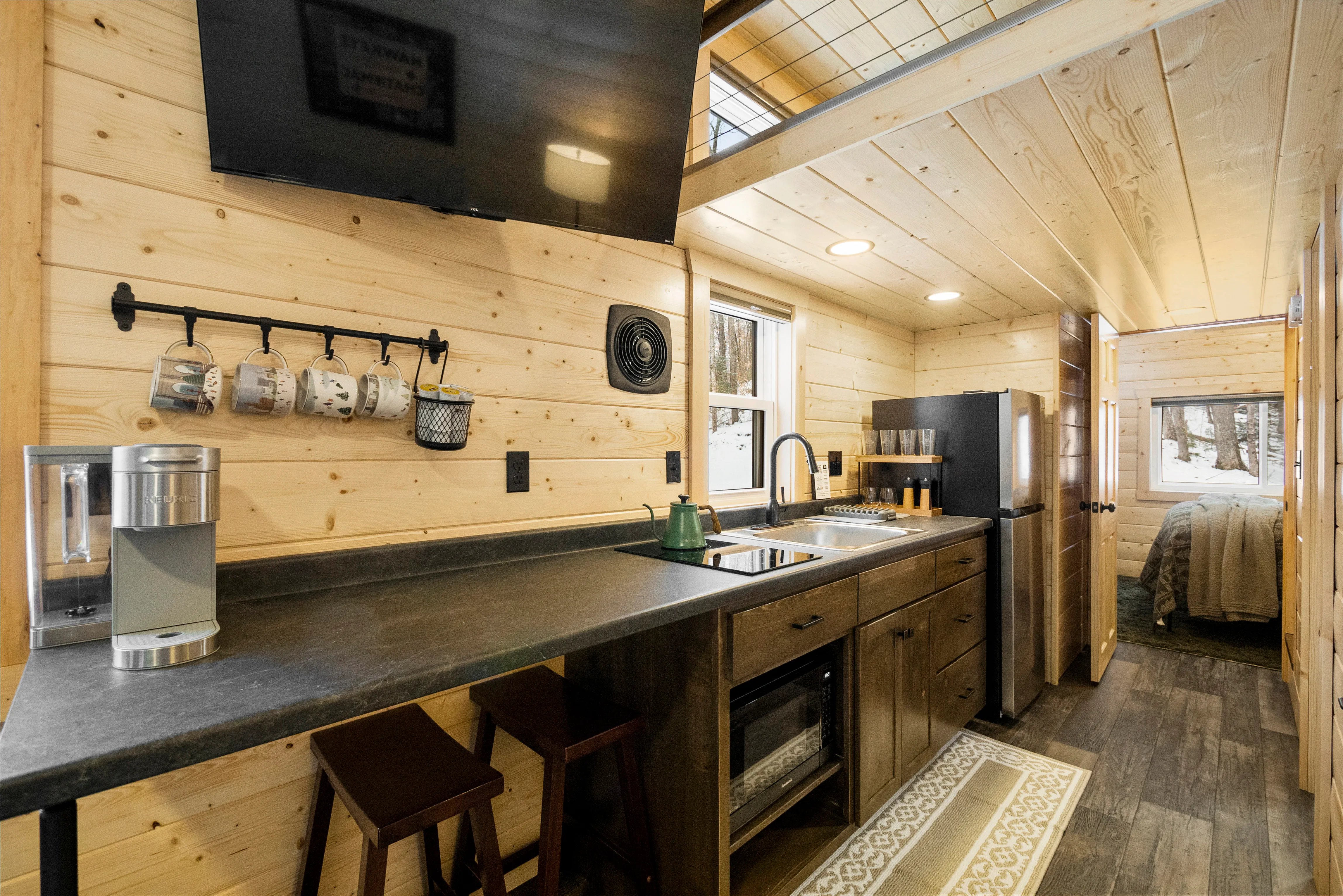 A compact kitchen with wood-paneled walls, black countertop, sink, stove, coffee maker, mugs, and a wall-mounted TV, leading to a bedroom at the end of the hallway.