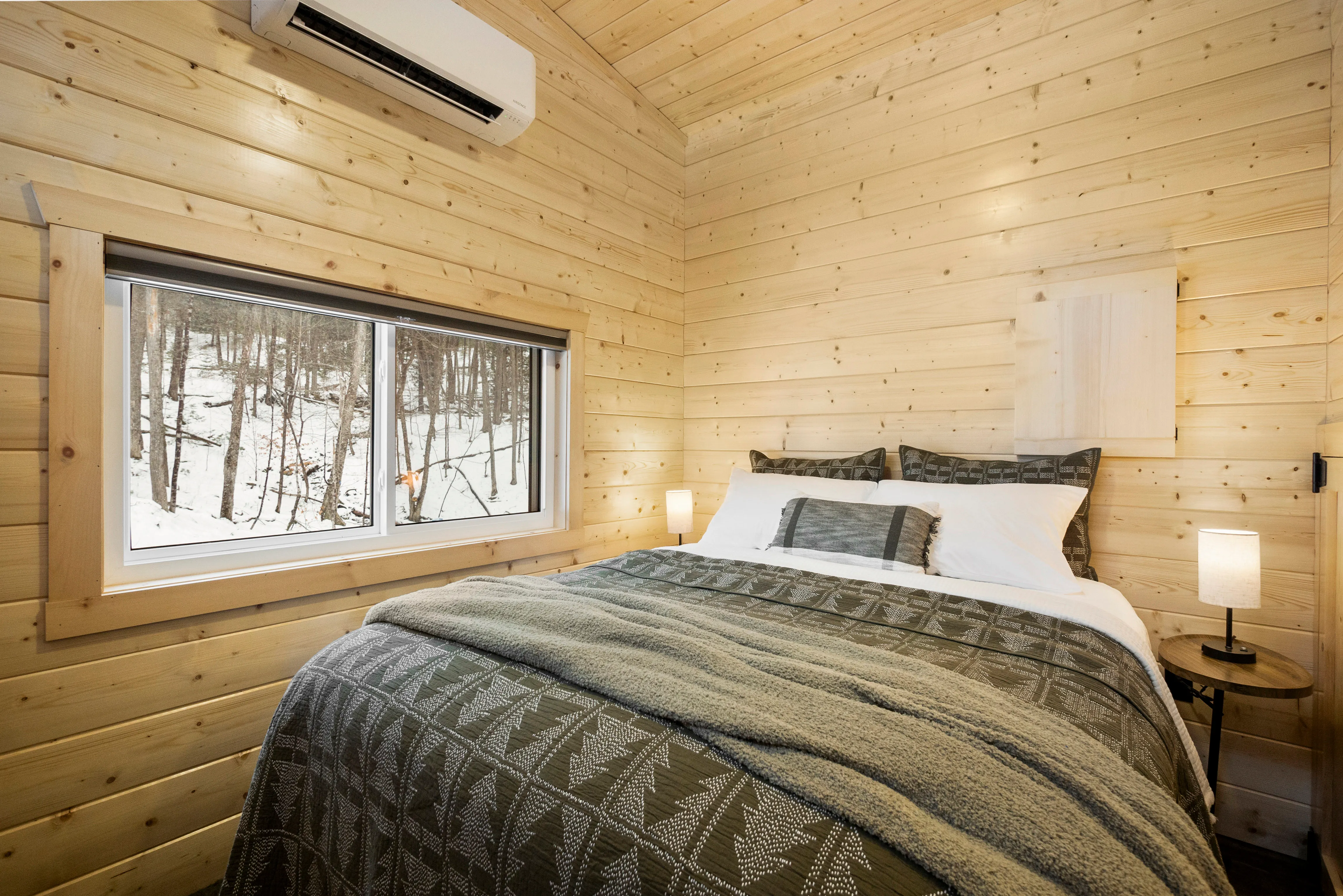 A small bedroom with wood-paneled walls, a double bed with green patterned bedding, two bedside tables with lamps, and a window.