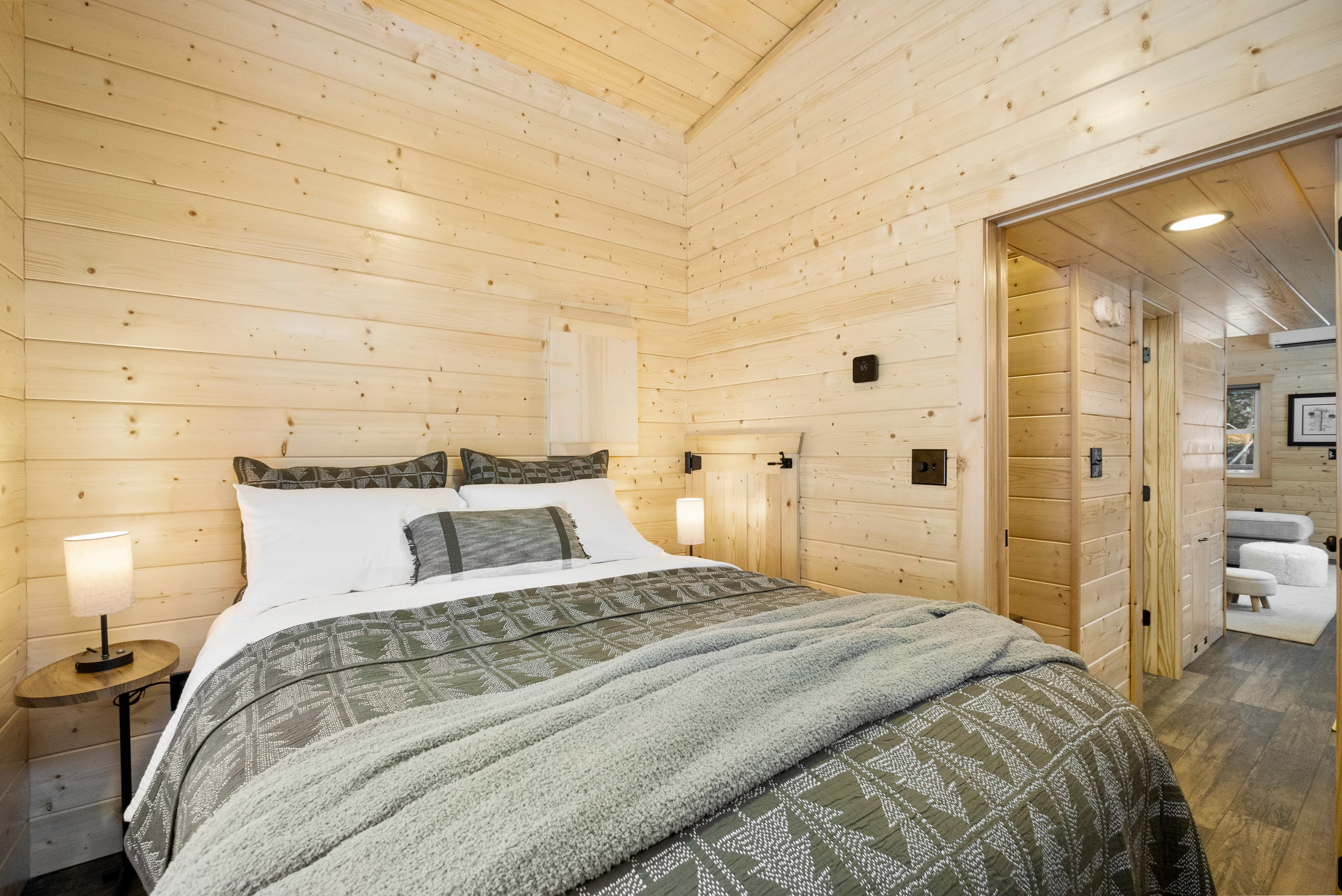 A cozy bedroom with light wood-paneled walls, a double bed with green patterned bedding, two side tables with lamps, and an open doorway leading to another room.