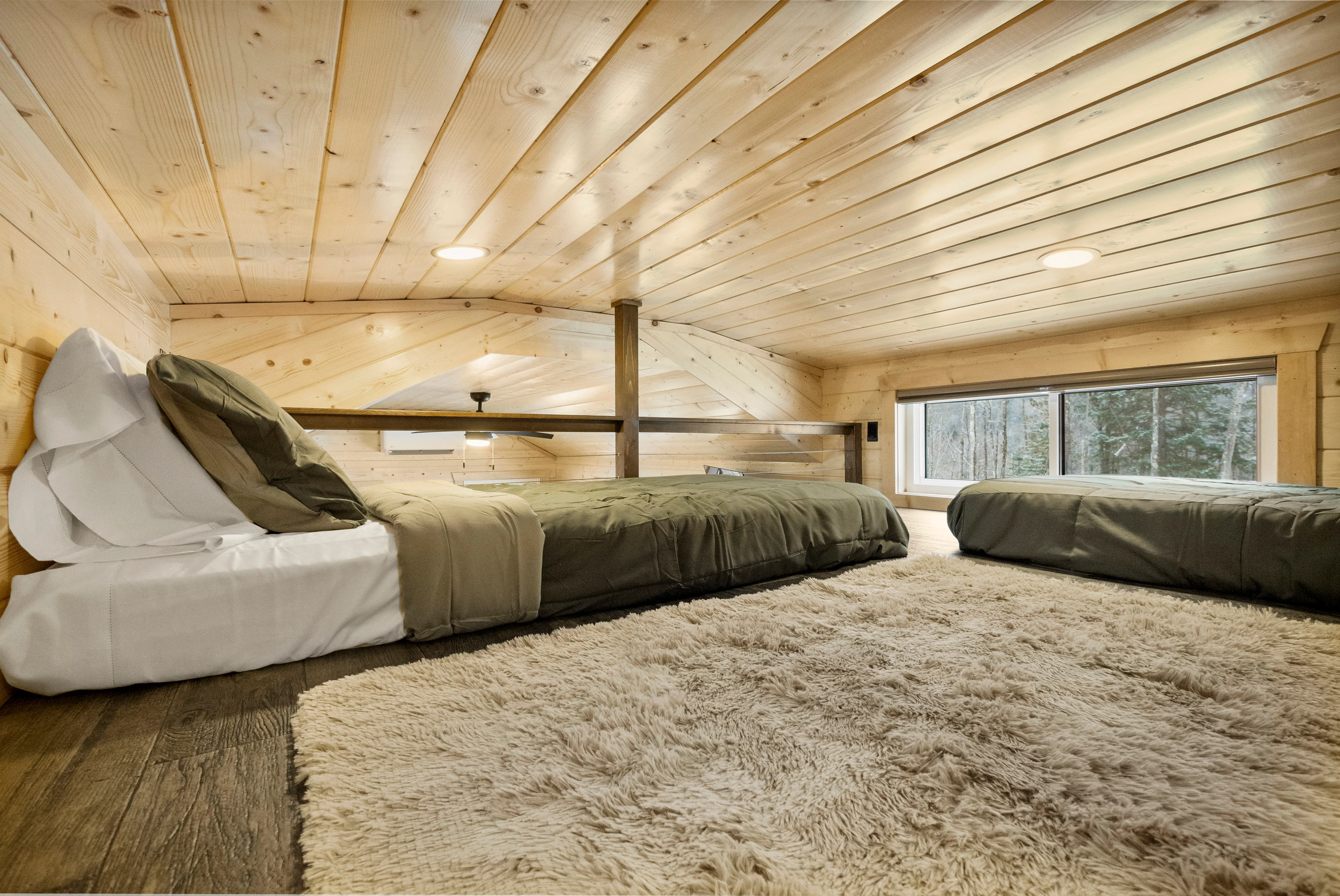 Two twin beds with green bedding on either side of a shaggy beige rug in a wood-paneled loft with a large window.