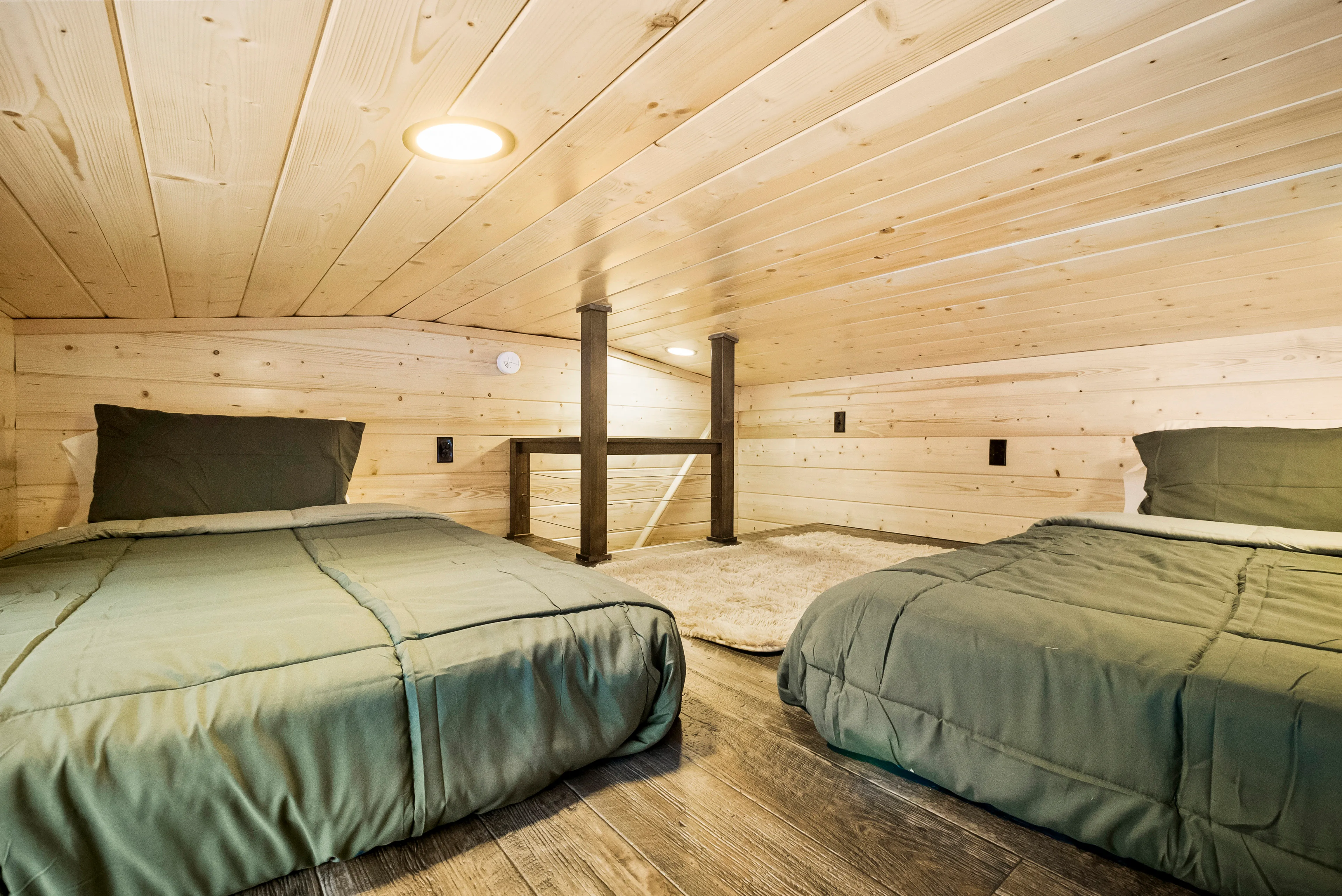 A small loft with wood-paneled walls and ceiling features two beds with green bedding, a beige rug, and a wooden floor.