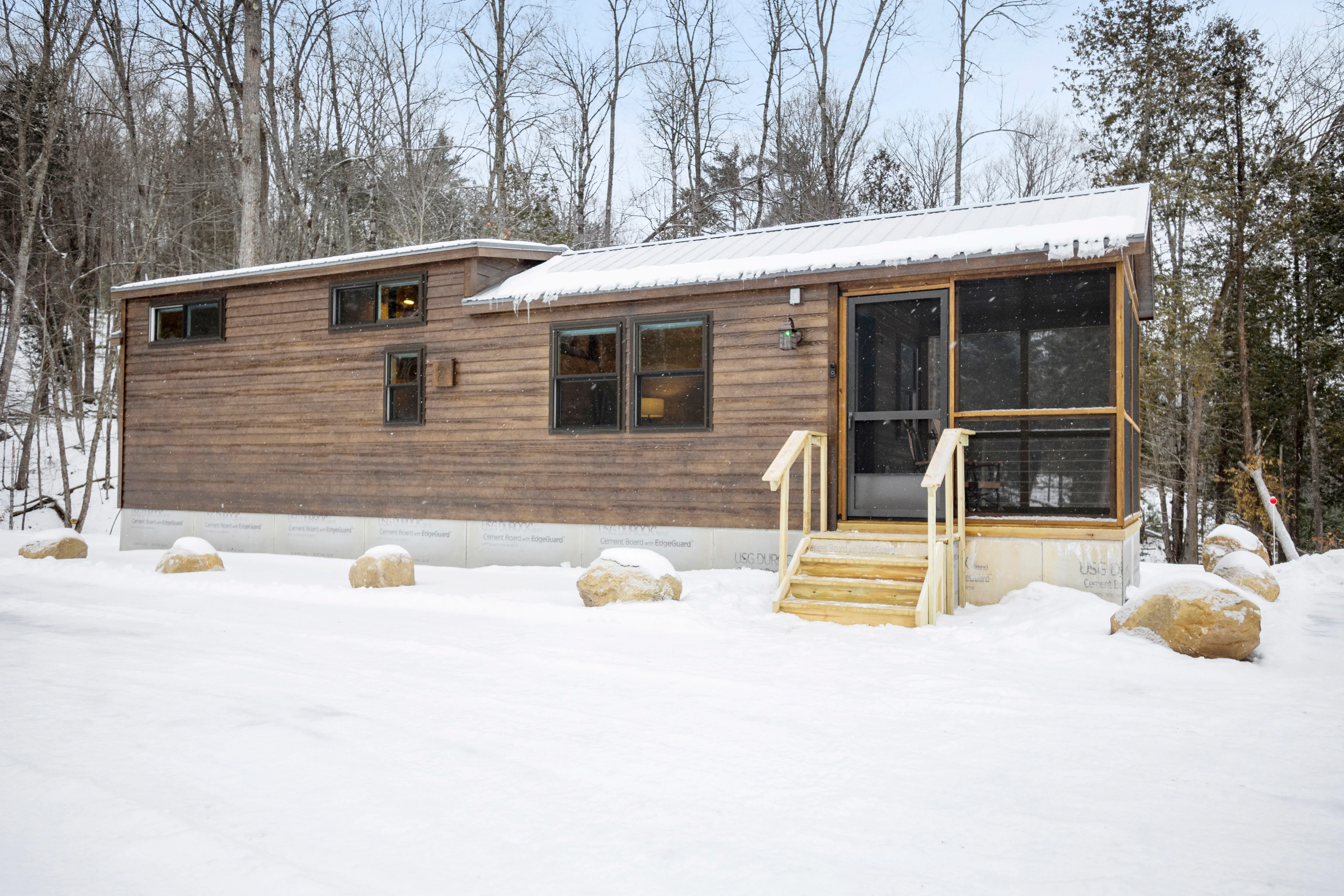 A small wooden house with a screened porch sits on a snowy lot surrounded by trees, with snow covering the roof and ground.