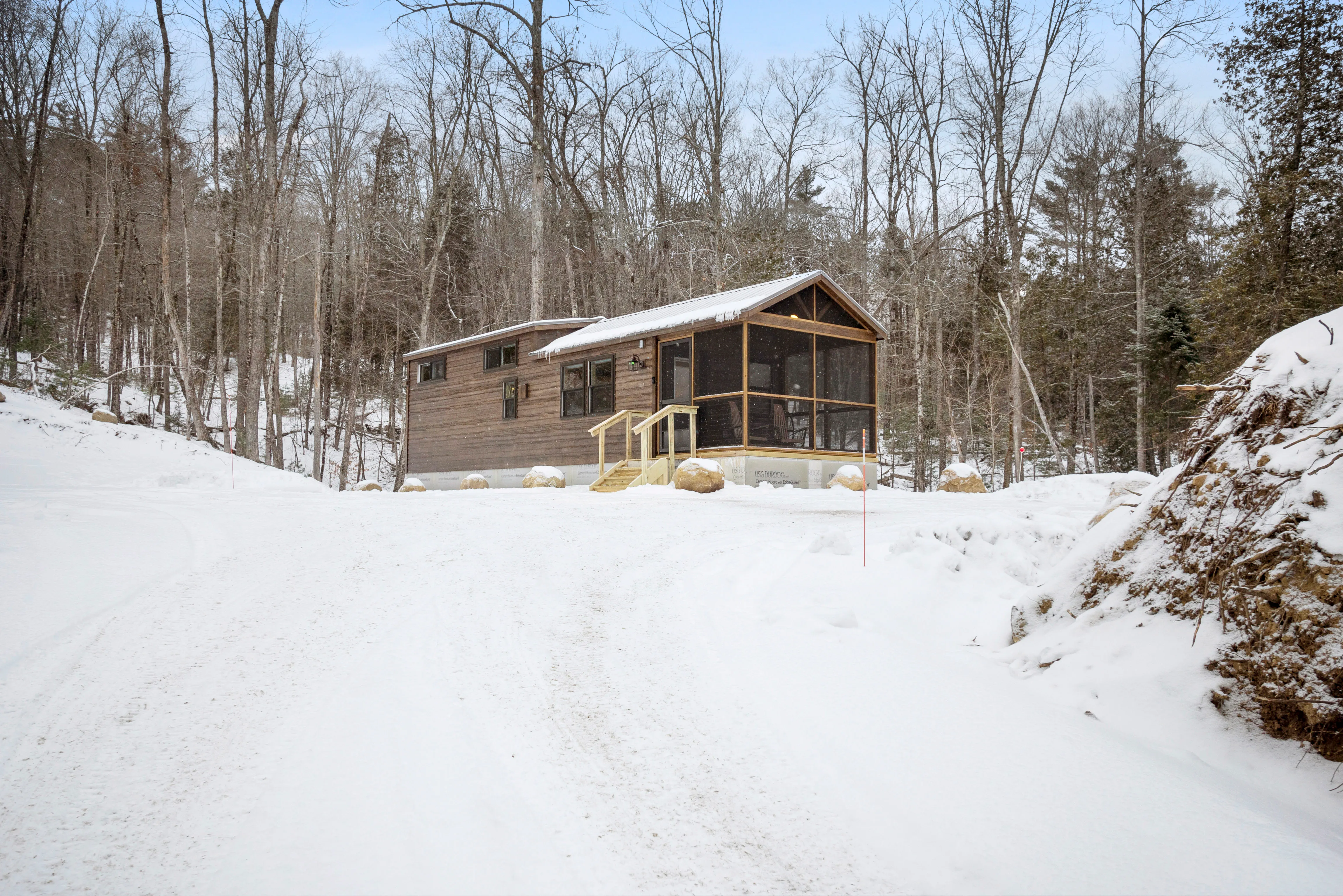 A small wooden cabin with a screened porch sits on a snowy hill surrounded by bare trees in a winter forest landscape.
