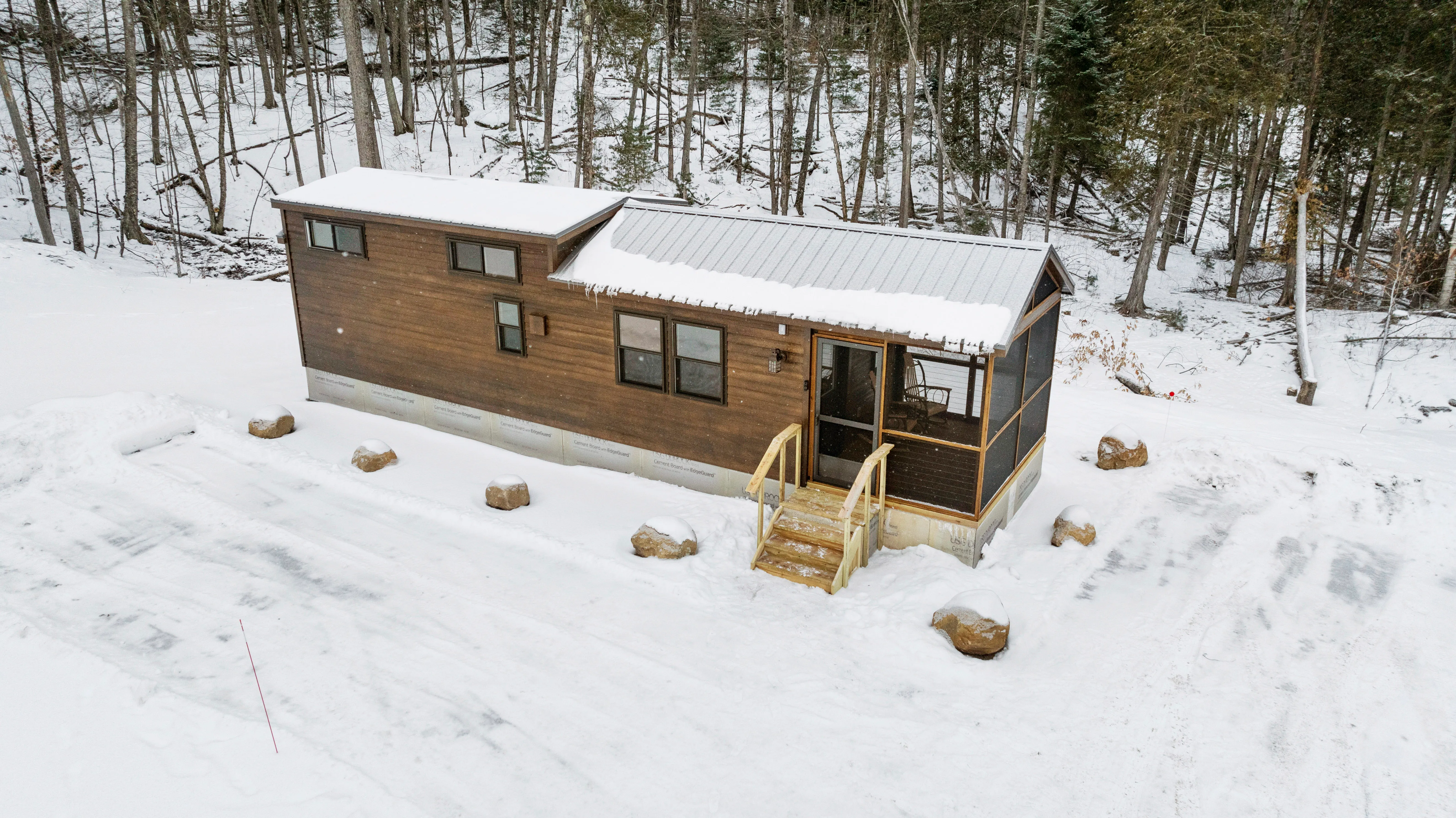A small wooden cabin with a metal roof sits in a snowy clearing surrounded by trees.