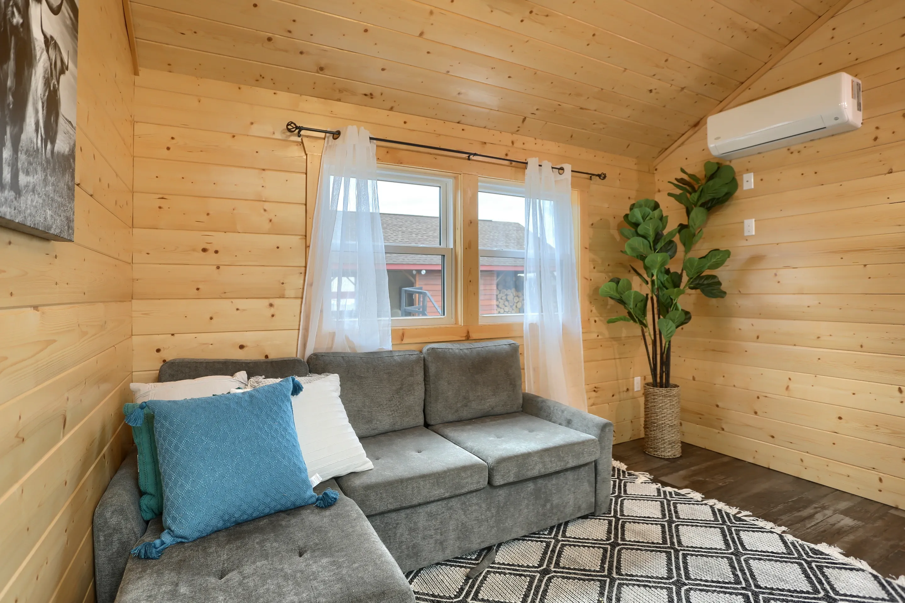 A small room with wood-paneled walls, a gray sectional sofa with cushions, a patterned rug, a potted plant, sheer curtains, and a wall-mounted air conditioner.