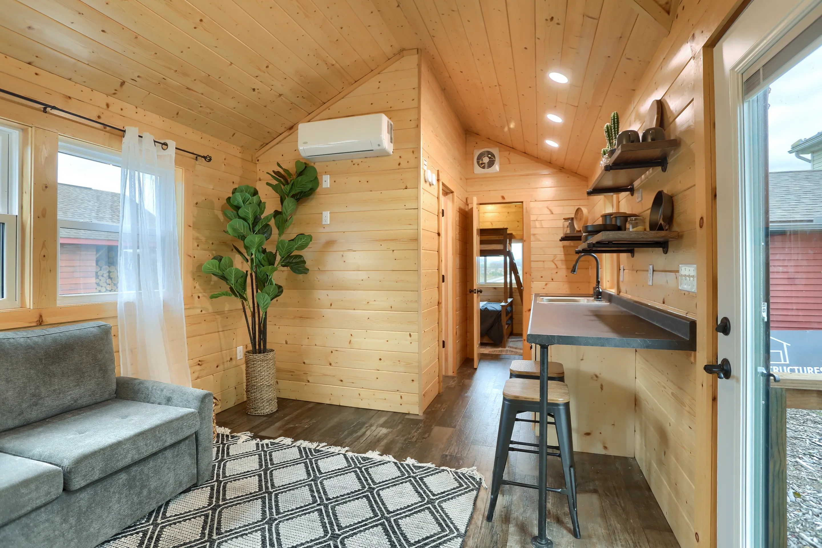 Interior of a small wooden cabin featuring a gray sofa, patterned rug, bar seating at a counter, potted plant, and open shelves with decor.
