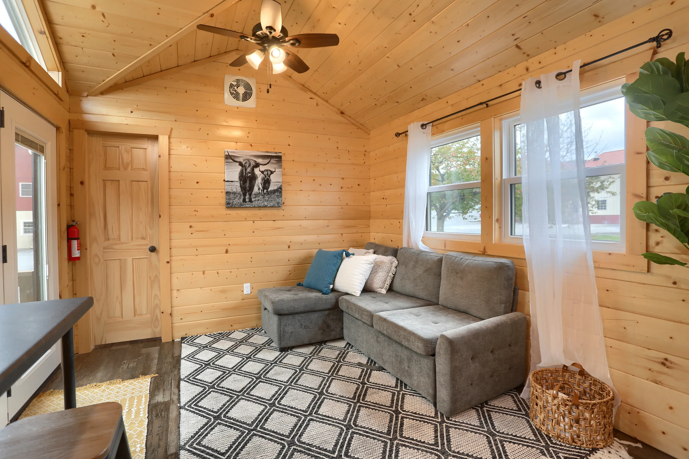 A small living room with wood-paneled walls, a gray sectional sofa with pillows, a patterned rug, wall art, large windows with white curtains, and a ceiling fan.