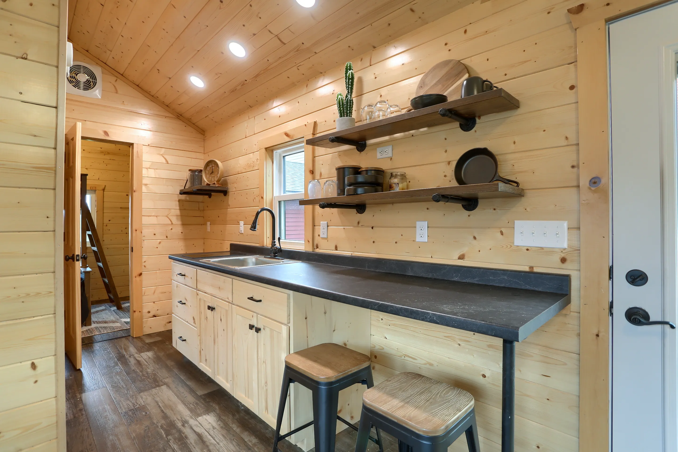 Modern kitchen with light wood walls, open shelves holding dishes, a black countertop with a sink, two wooden stools, and recessed ceiling lights.