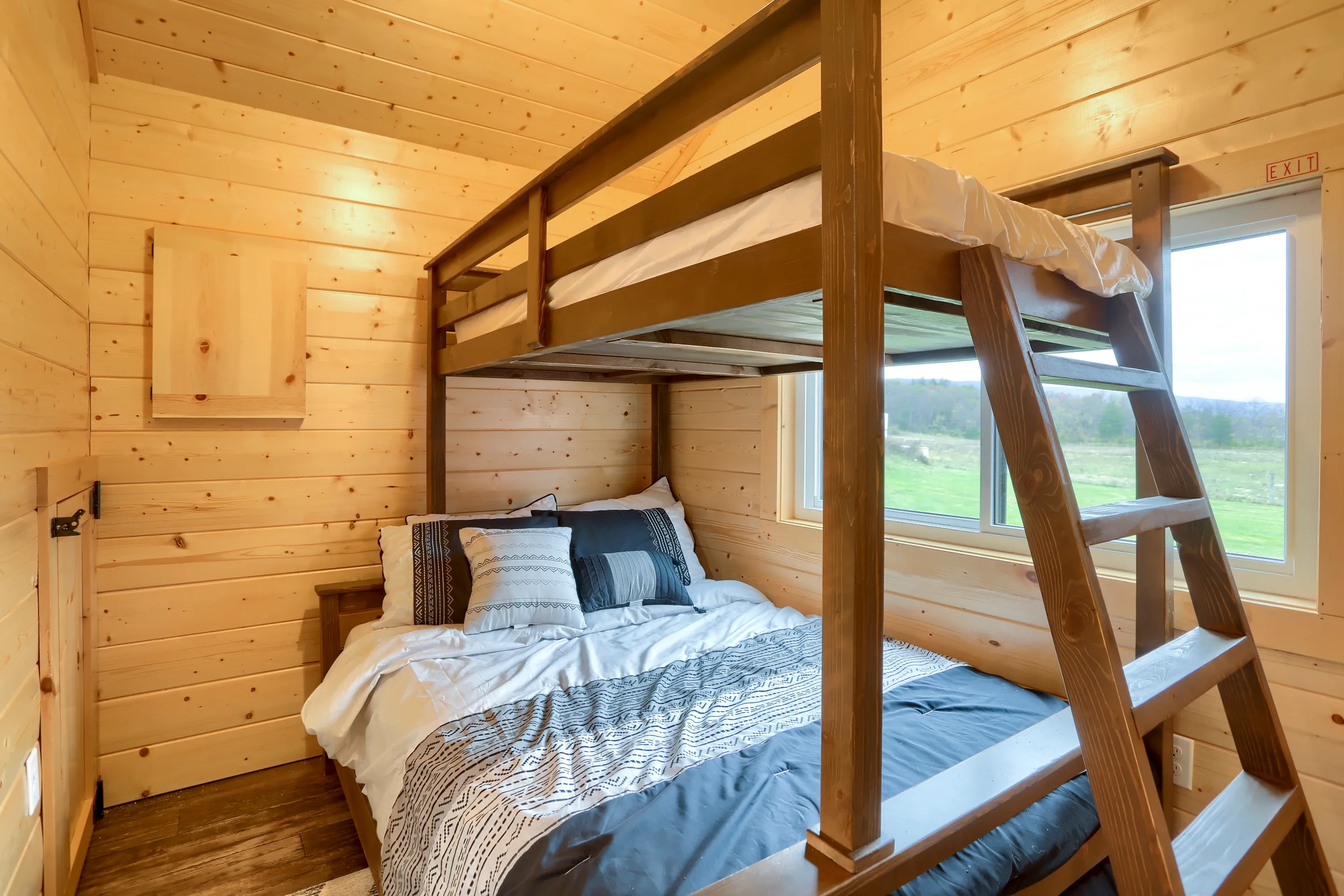A small wooden room with a double bed below and a single loft bed above, accessible by a wooden ladder, next to a window with an outdoor view.
