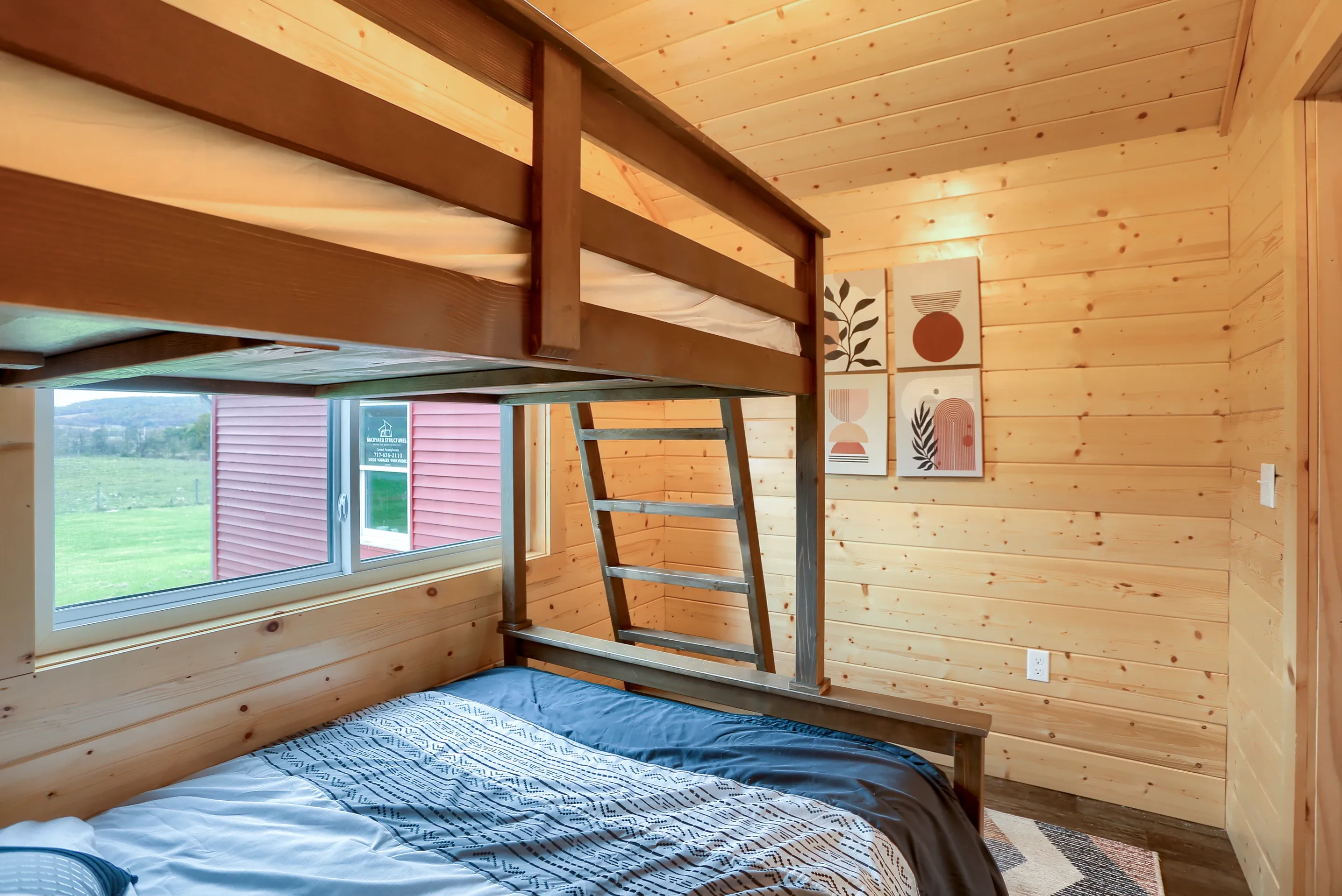 Small wooden room with double bed below and a single loft bed above, accessible by a wooden ladder, a window, a patterned bedspread, and minimalist artwork on the wall.