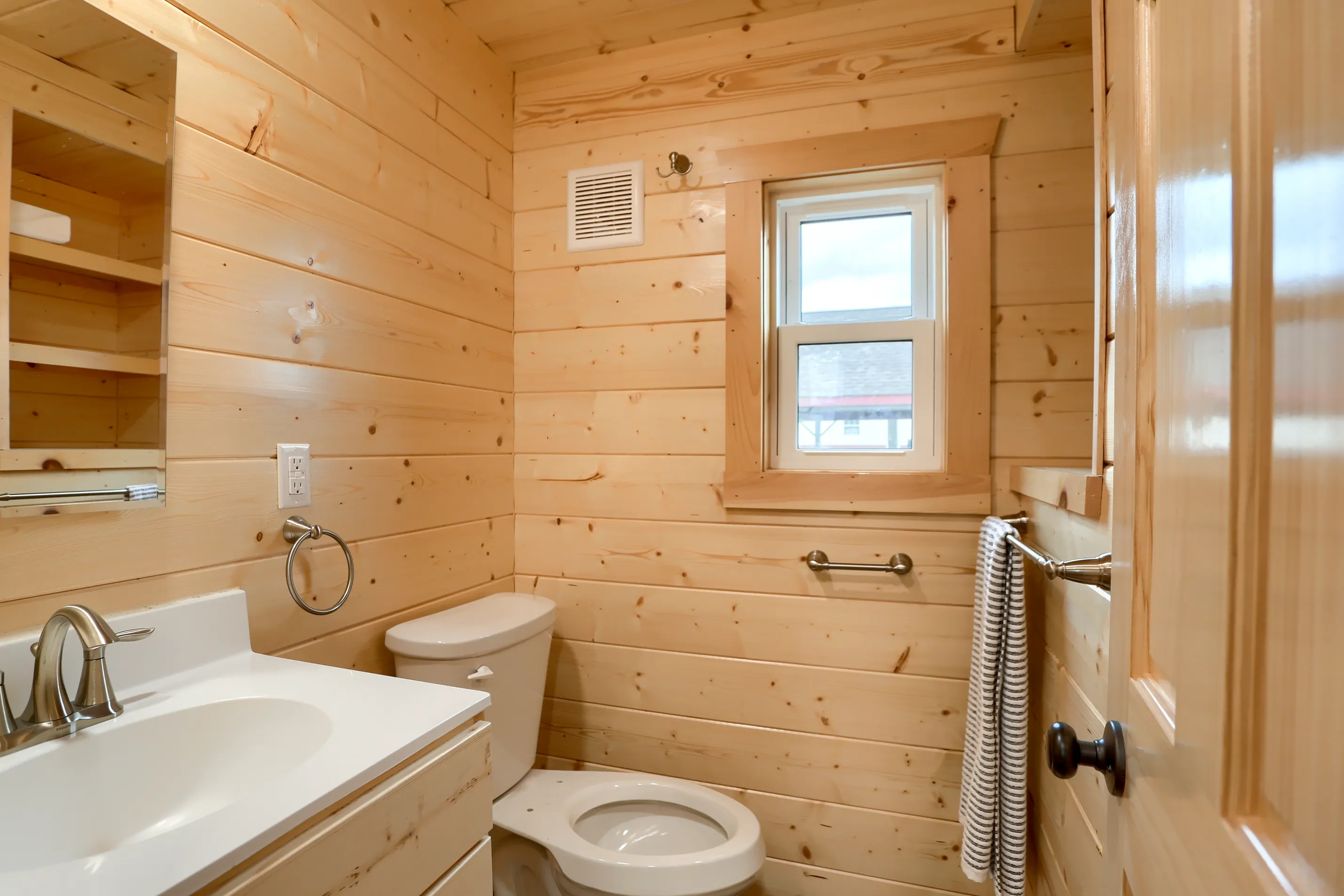 Small bathroom with light wood paneling, a white sink and toilet, a window, a mirror, and a towel hanging on a metal bar.