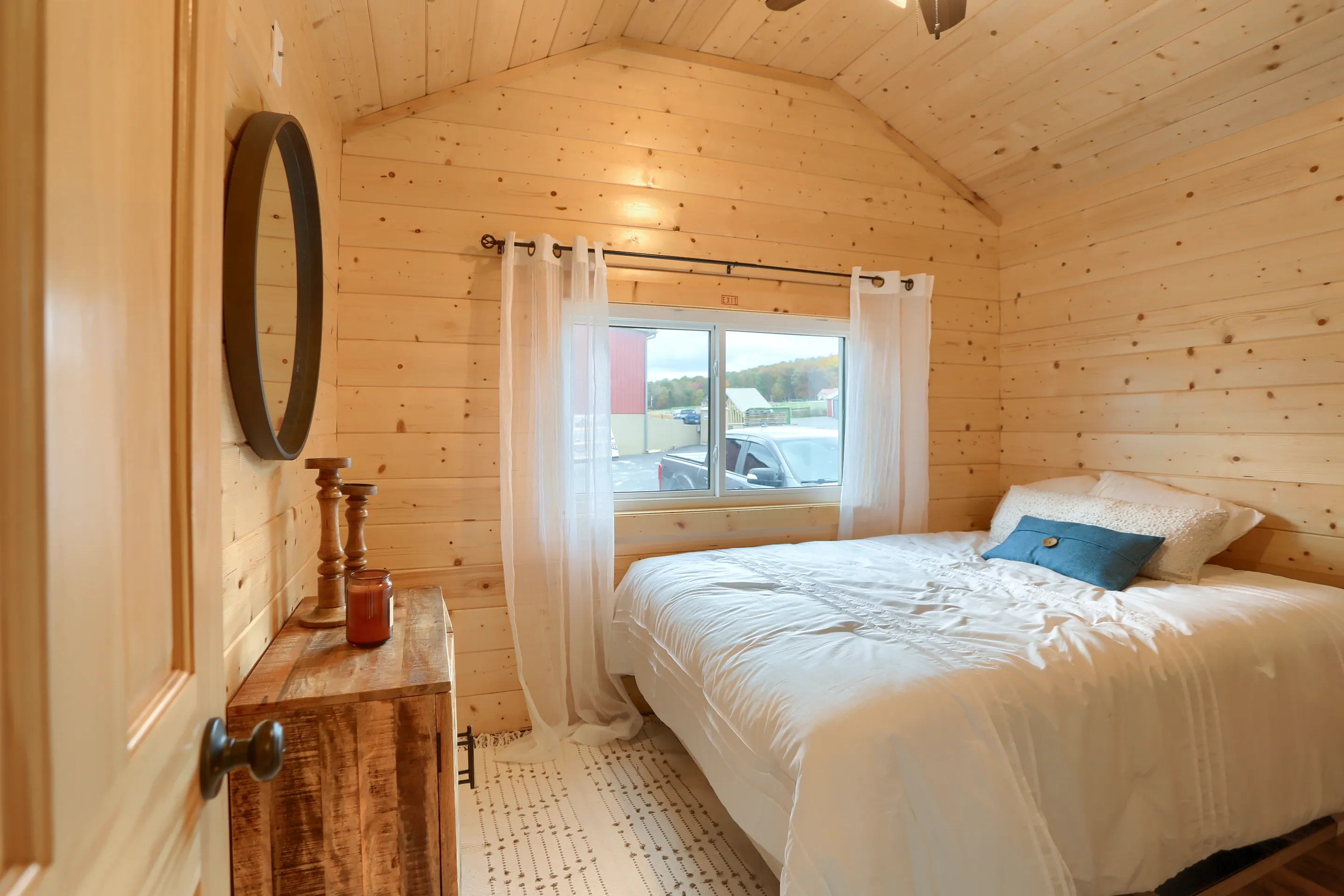 Small bedroom with light wood-paneled walls, a double bed with white bedding, a blue pillow, sheer curtains, a window, a wooden dresser and a circular mirror.