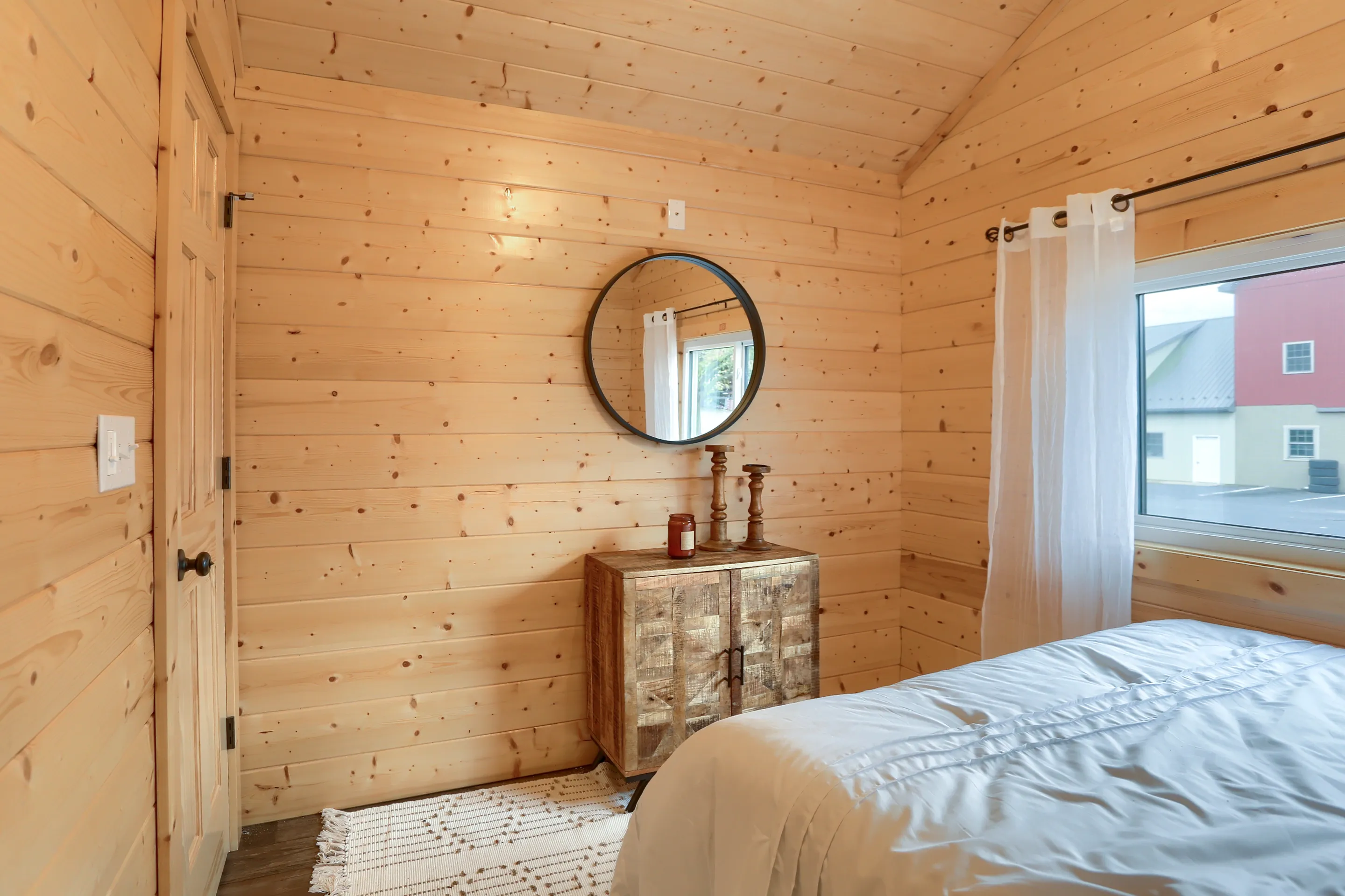 Bedroom with light wood-paneled walls, a round mirror above a wooden cabinet, white curtains, and part of a bed visible in the foreground.