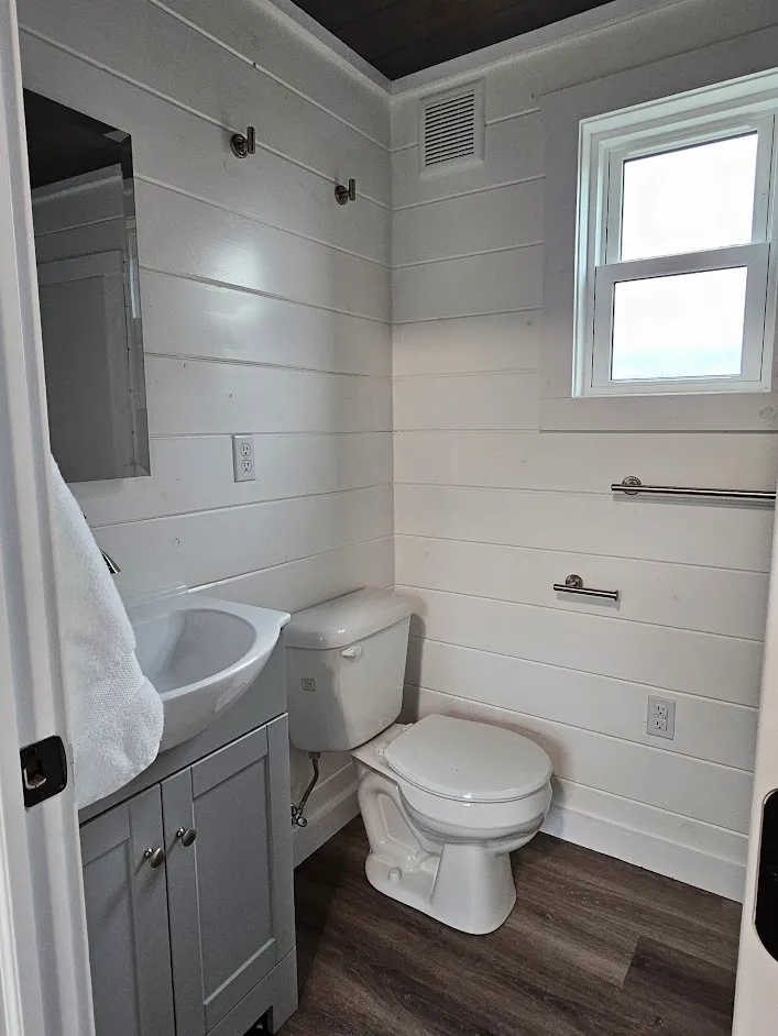 Small bathroom with white shiplap walls, a window, a toilet, a gray vanity with sink, towel hook, and towel bar. The floor is wood-patterned.