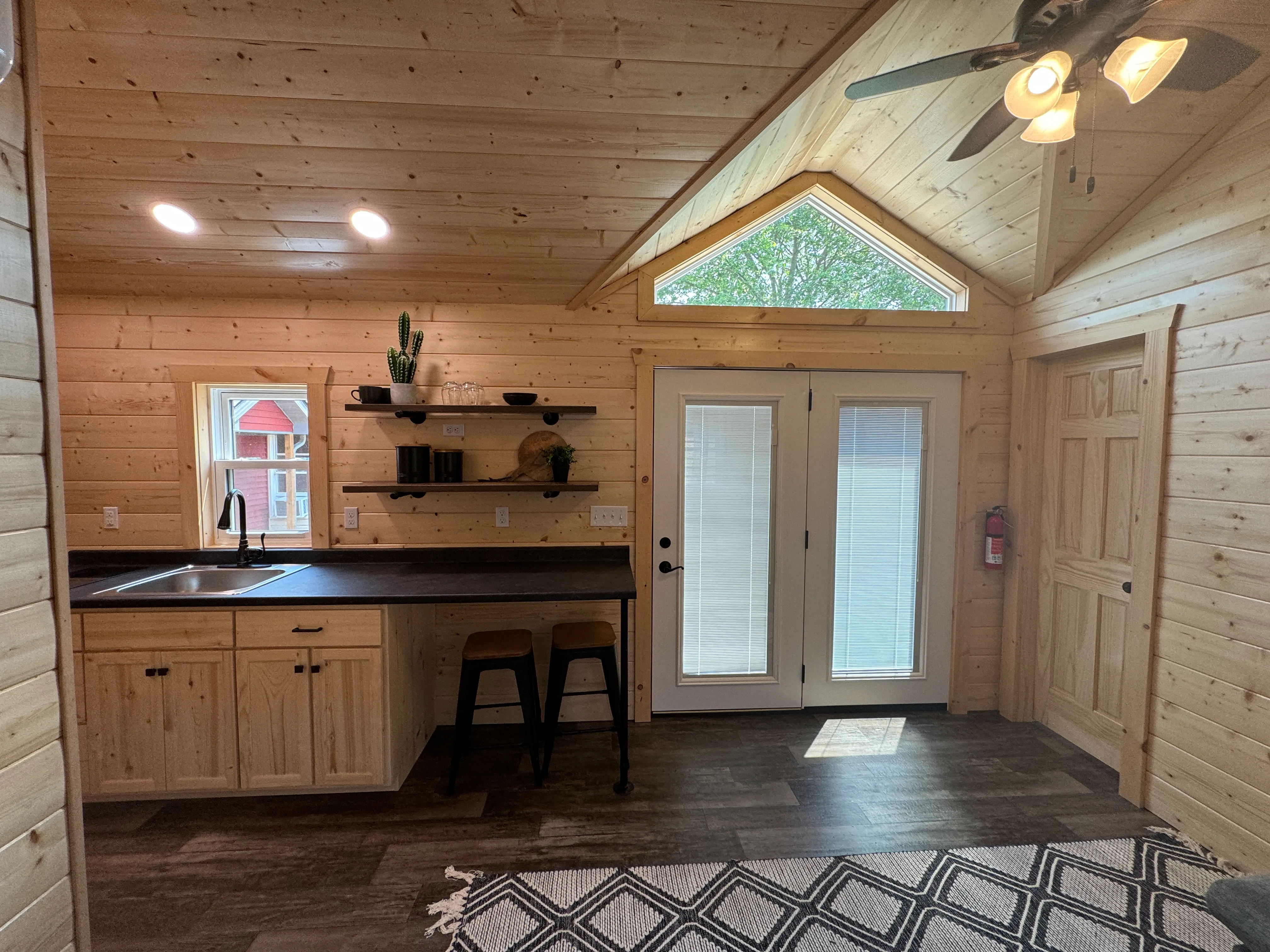 A compact kitchen with wooden cabinets, black countertop, open shelves, two stools, and double glass doors under a triangular window in a wood-paneled room.