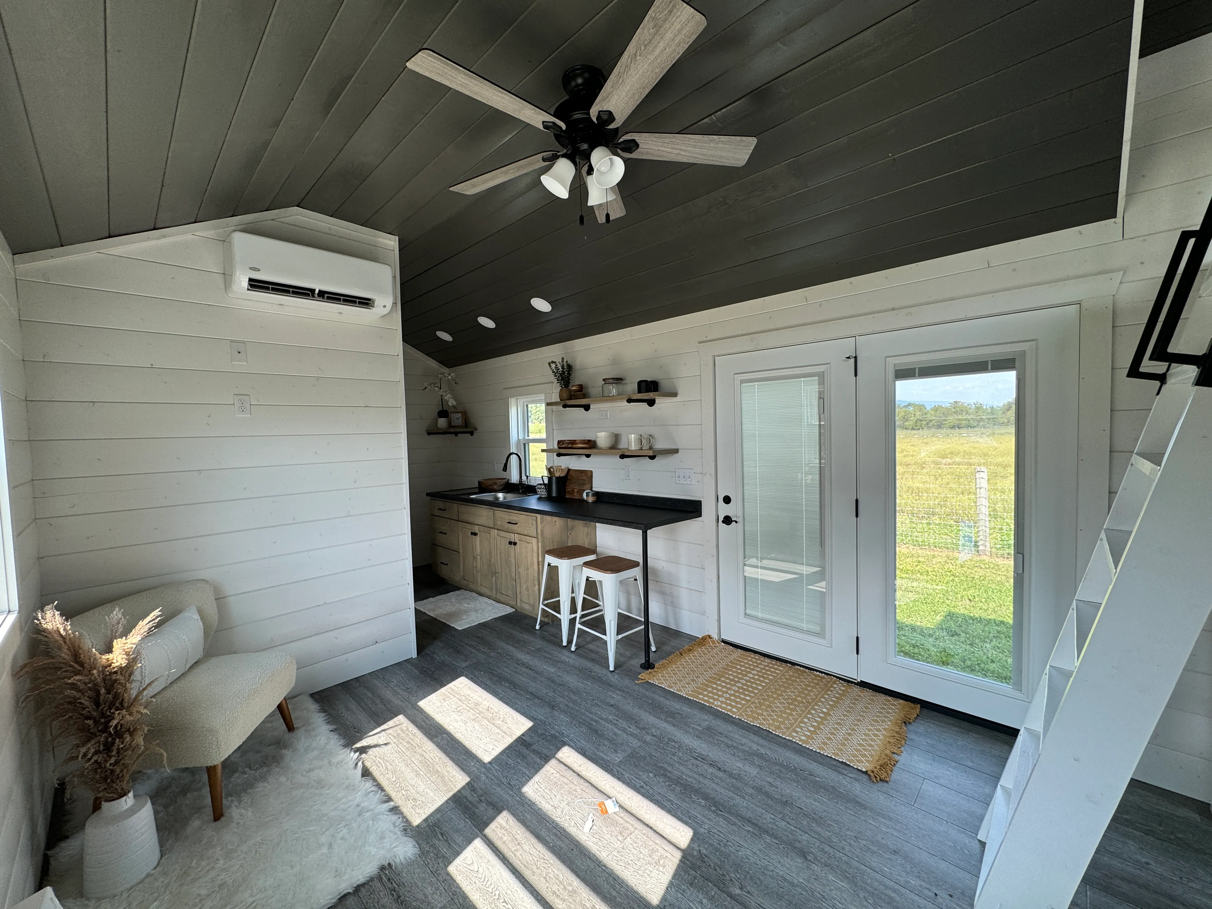 A modern tiny house interior with a kitchenette, bar stools, armchair, and loft ladder. Large windows and glass doors.