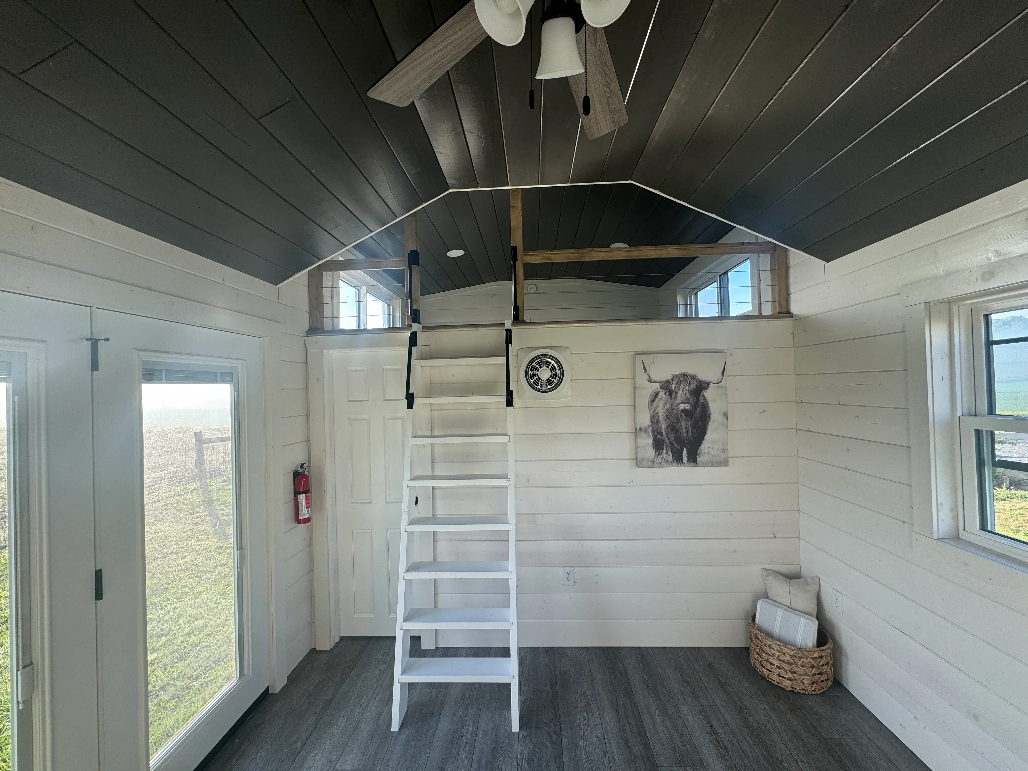 Interior of a small room with white wood walls, a loft with ladder stairs, a ceiling fan, a wall clock, and a basket with towels.