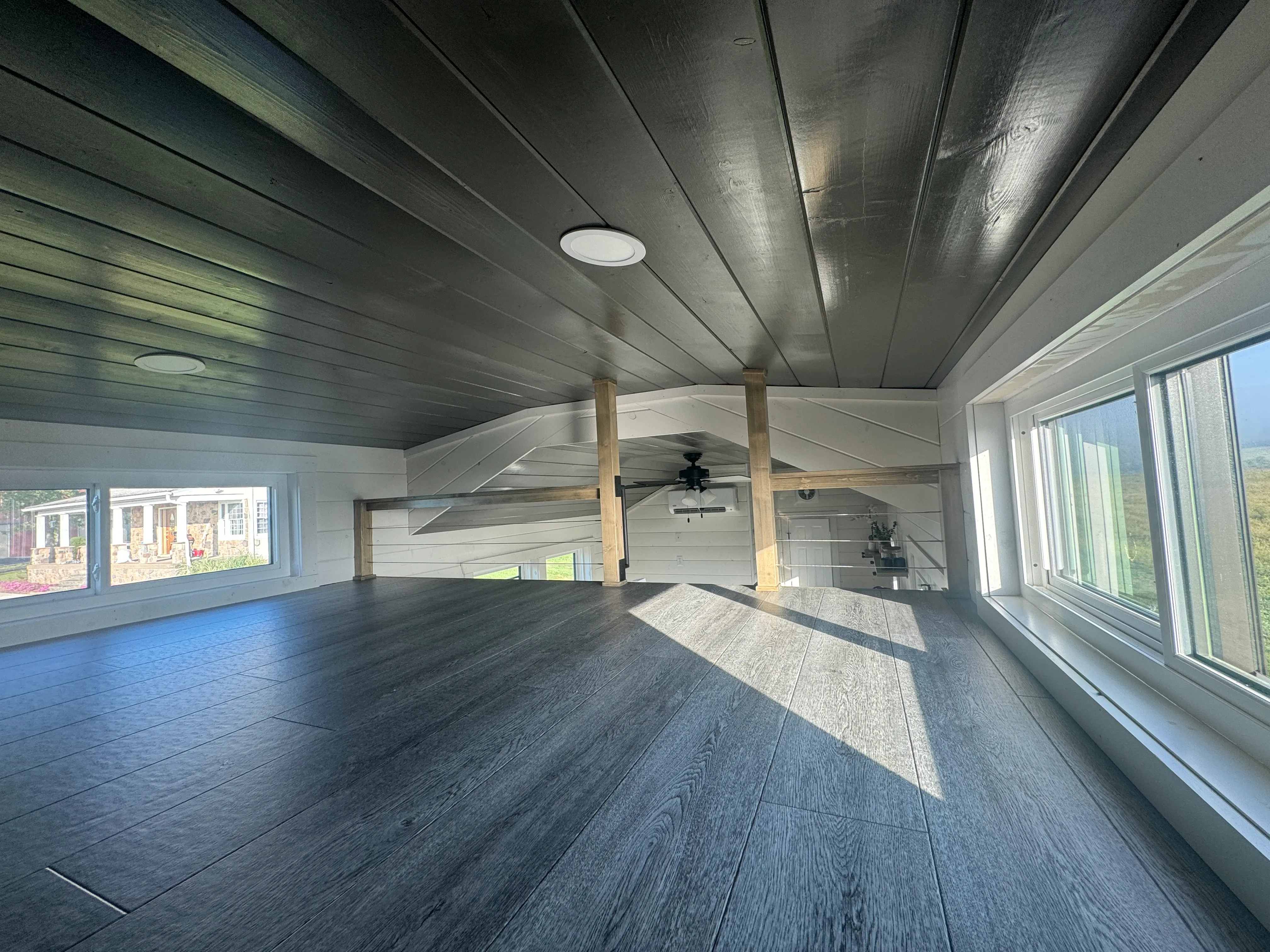 A small loft space with dark wood floors and ceiling, white walls, large windows, and exposed wooden beams, overlooking the area below.