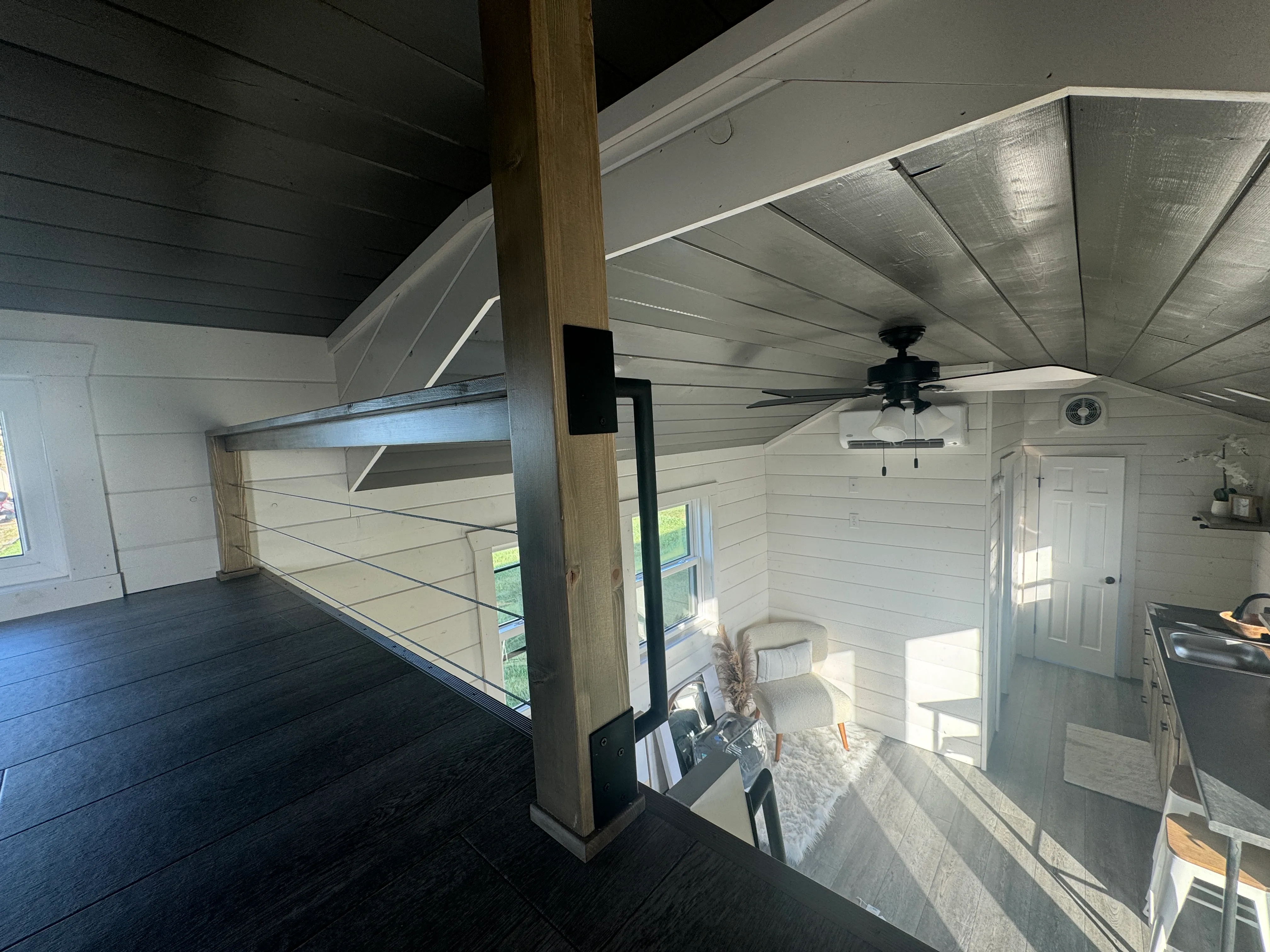 Interior of a small modern home with a loft area, white walls, dark flooring, ceiling fan, and a compact kitchen and living space below.